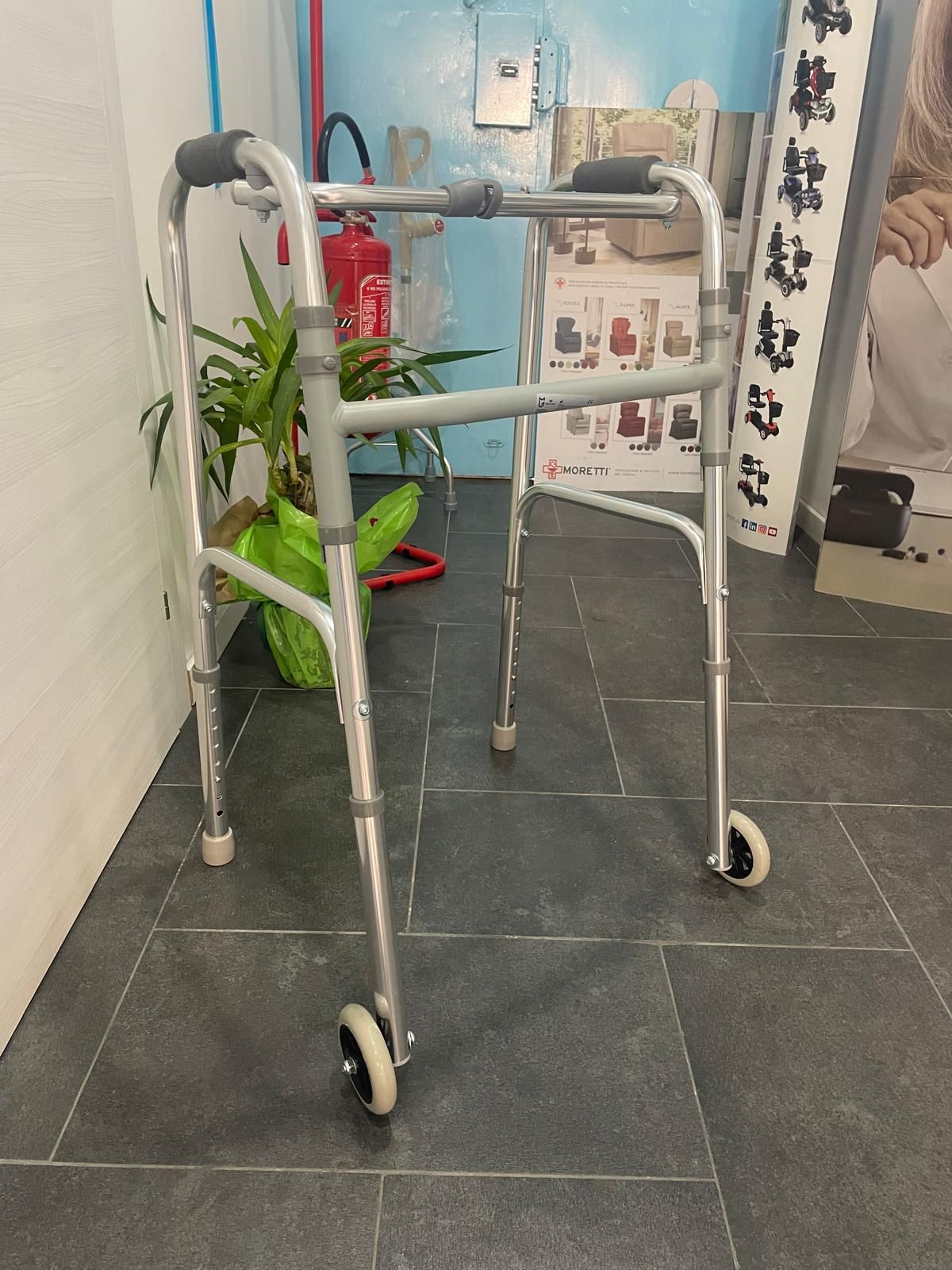 Deambulatore argentato con ruote, in casa, vicino a una pianta e a un estintore.