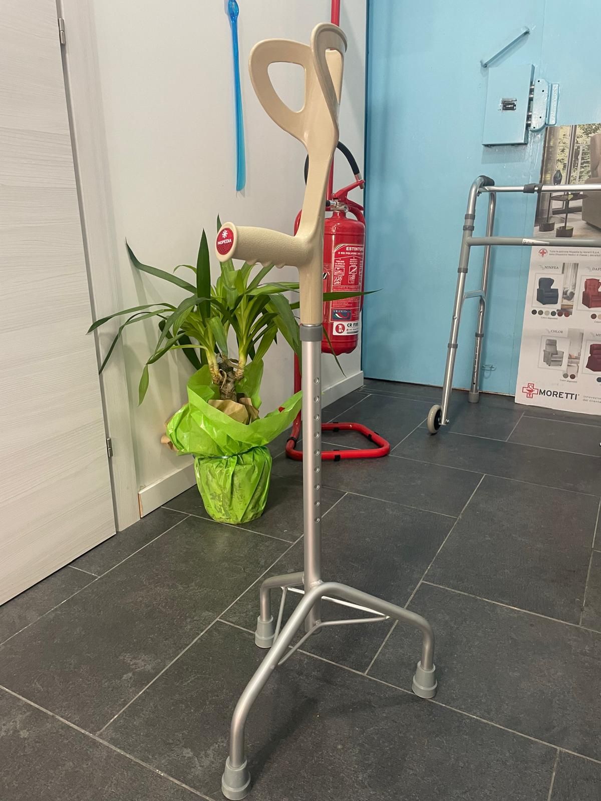 Un bastone quadrato, grigio e beige, è appoggiato su un pavimento piastrellato, vicino a una pianta e a un muro.