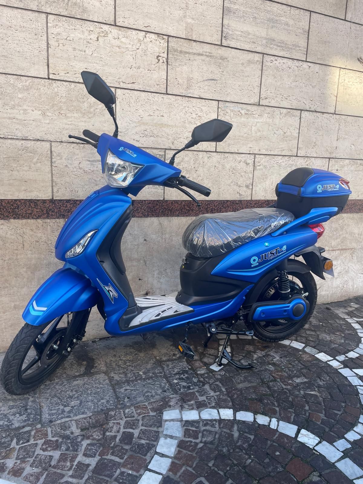 Monopattino elettrico blu parcheggiato contro un muro di mattoni. Contenitore nero sul retro.