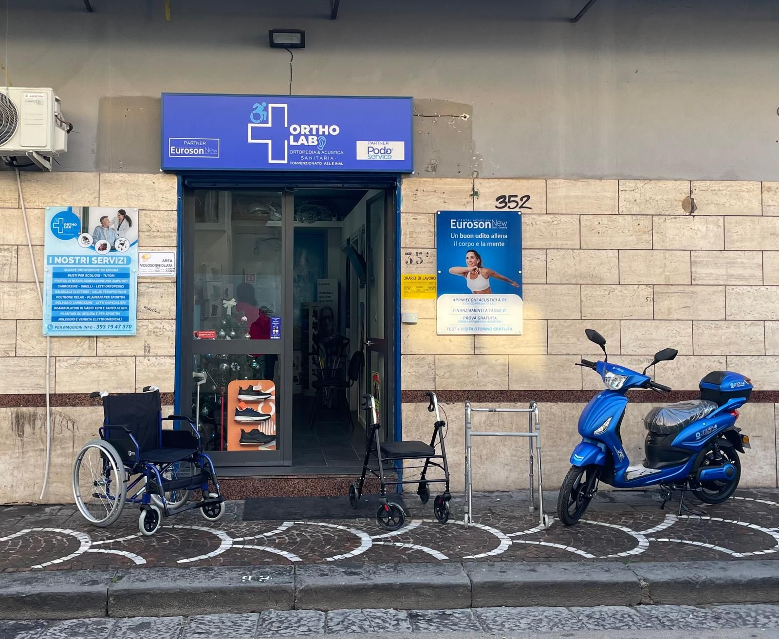 Esterno di un negozio di ortopedia con una sedia a rotelle, un deambulatore, uno scooter e un'insegna blu.