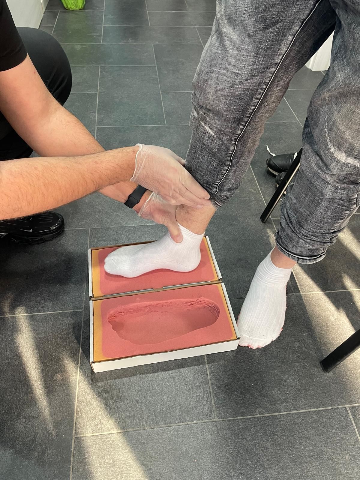 Piede di una persona in uno stampo, che riceve assistenza. Mani che indossano guanti. L'ambiente è al chiuso.