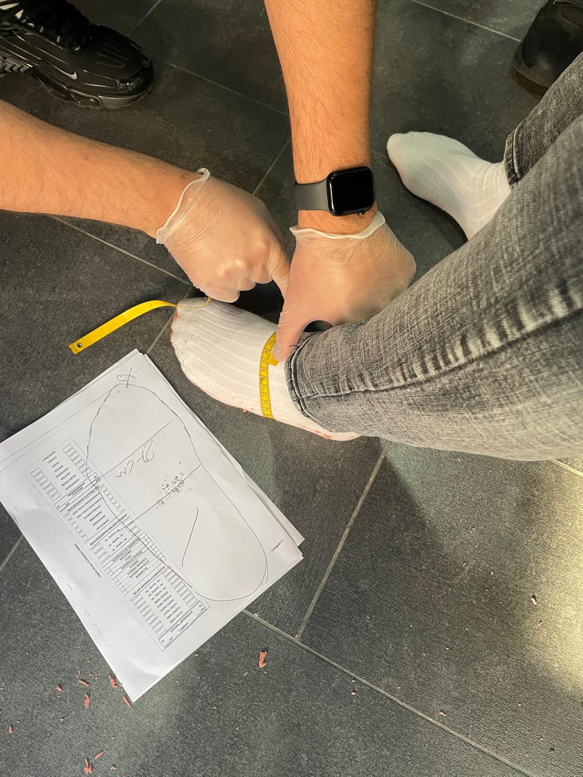 Due persone, sedute sul pavimento, indossano calzini bianchi e guanti e misurano un piede con un metro a nastro.