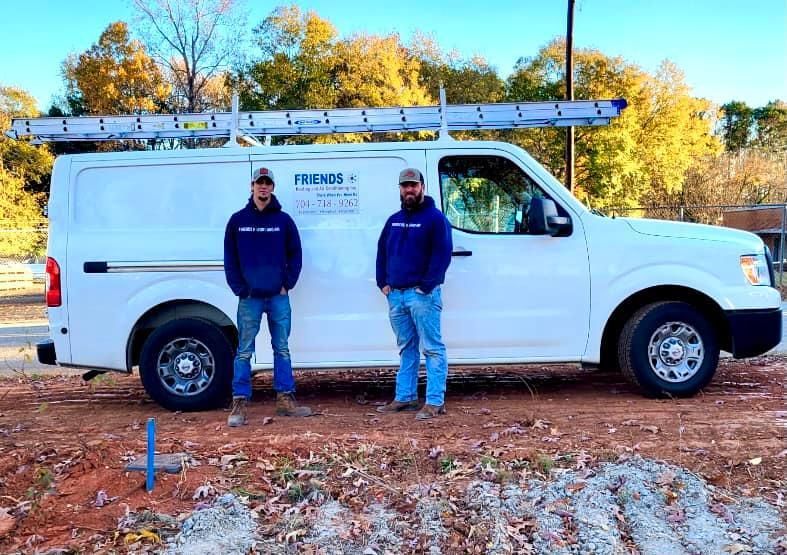 Two people stand in front of a white work van with a ladder on the roof on a dirt lot with trees in the background.