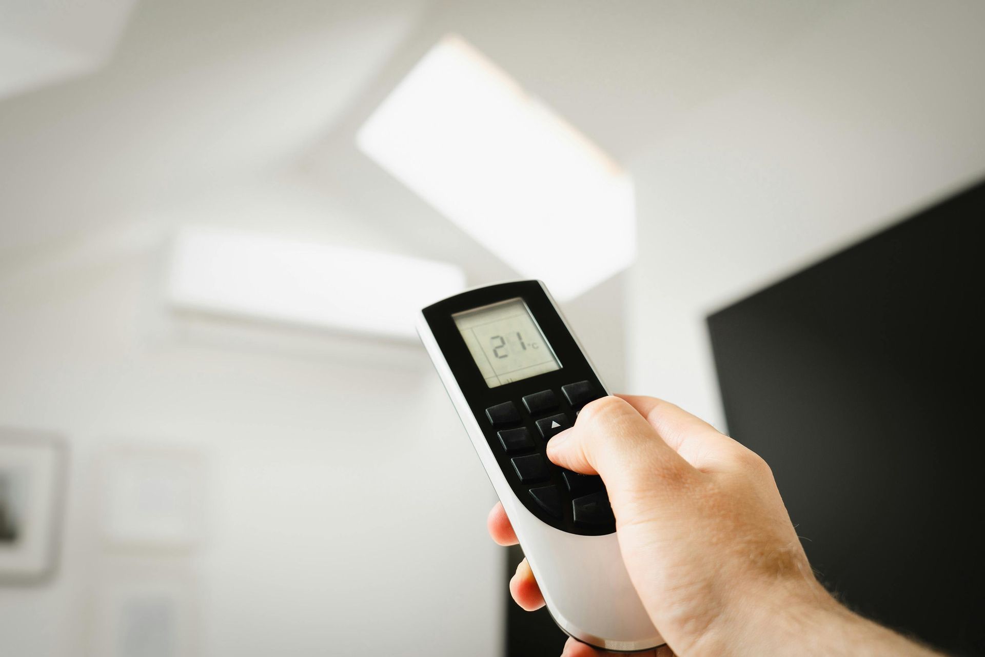 A hand holds a white remote control, pointing it toward a wall-mounted air conditioner in a bright room.