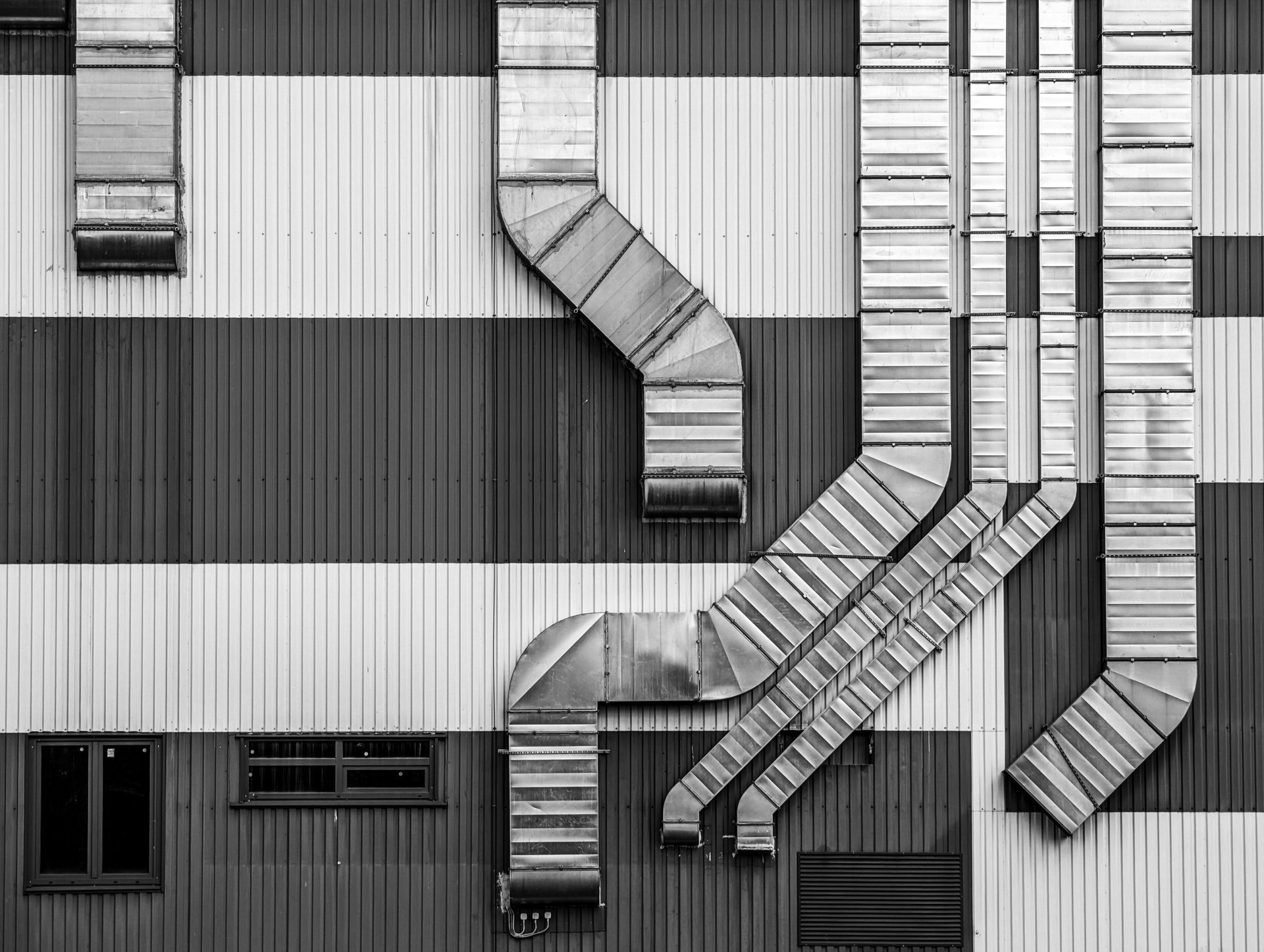 Industrial building wall with vertical metal siding, small windows, and complex metallic ventilation ductwork.