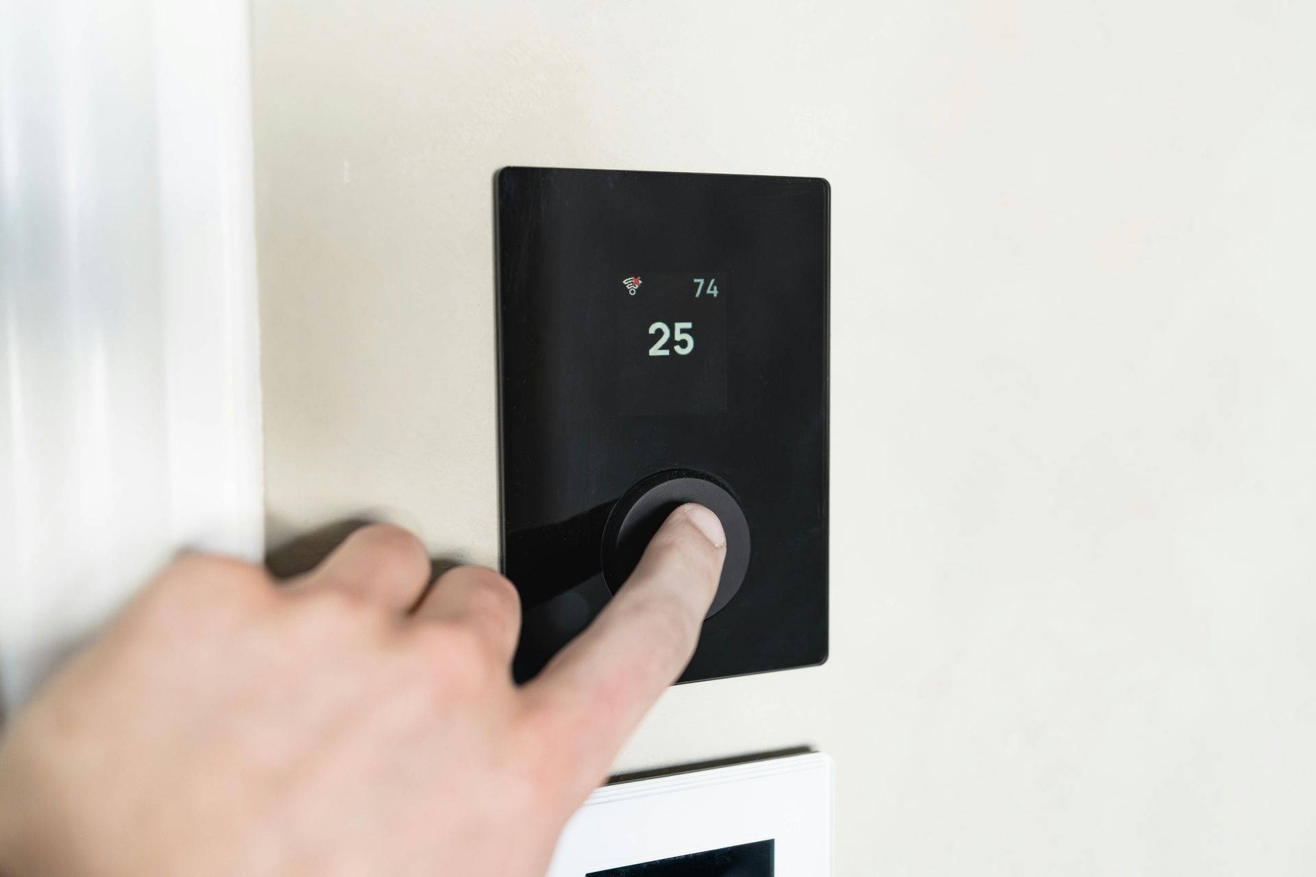 A hand presses a button on a sleek, black digital thermostat mounted on a light-colored wall.