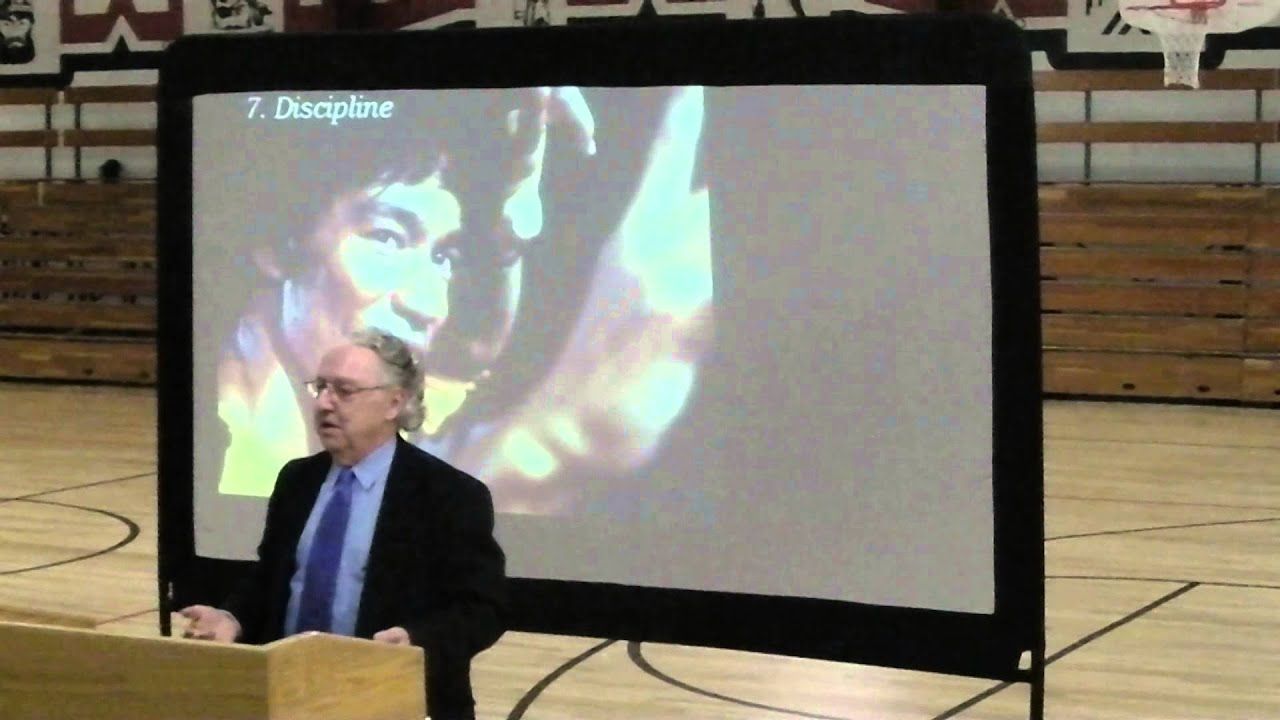 A man in a suit gives a presentation at a podium in a gymnasium, with an image of Bruce Lee on a projector screen.