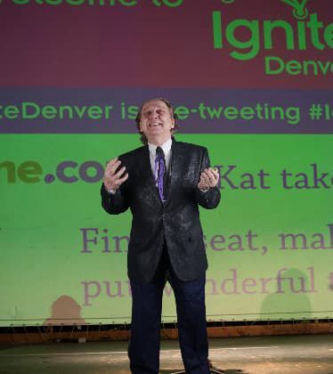 A person in a dark suit speaks expressively on stage in front of a large, green projected screen at Ignite Denver.