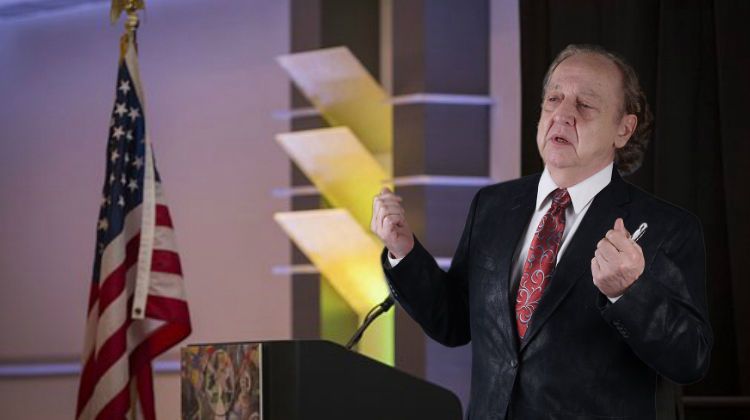 A person in a suit speaks at a podium next to an American flag, gesturing with their hands.