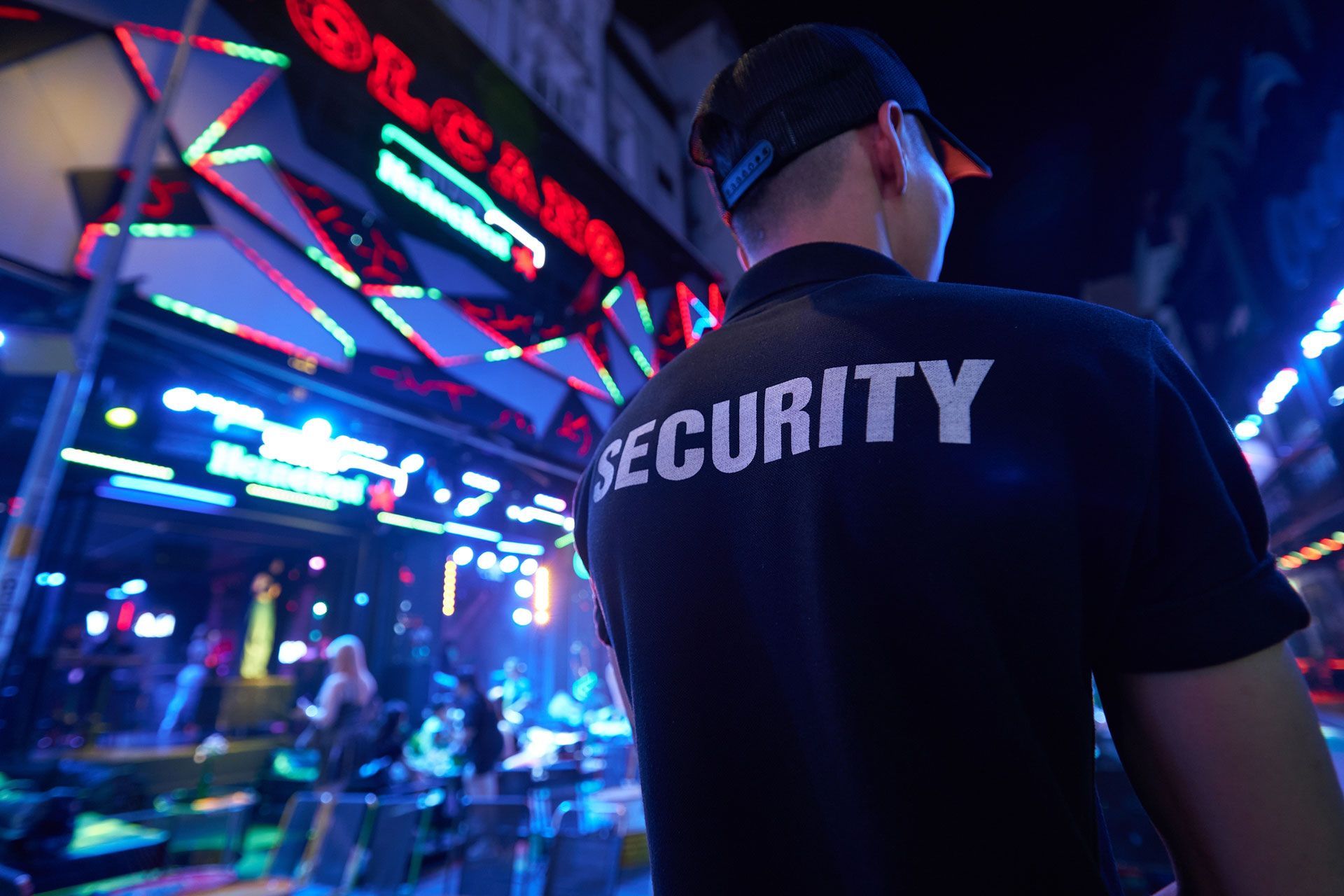 Security guard in black uniform stands outside a brightly lit nightclub.