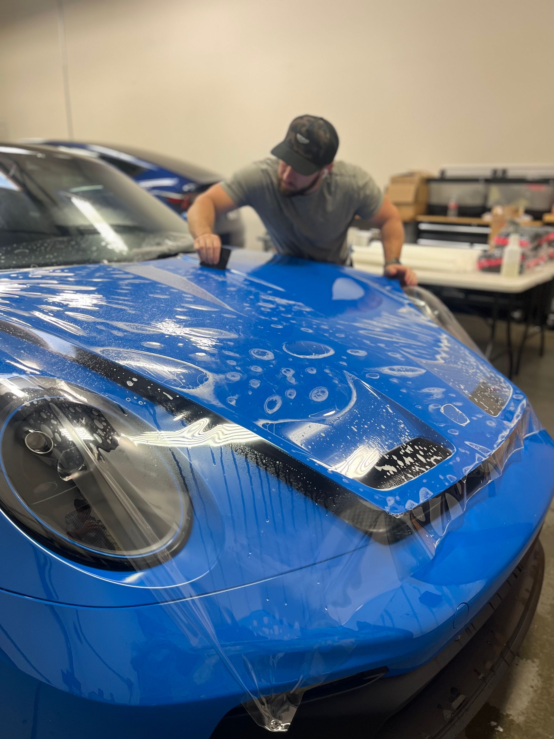 A man is covering the hood of a blue car with plastic wrap.