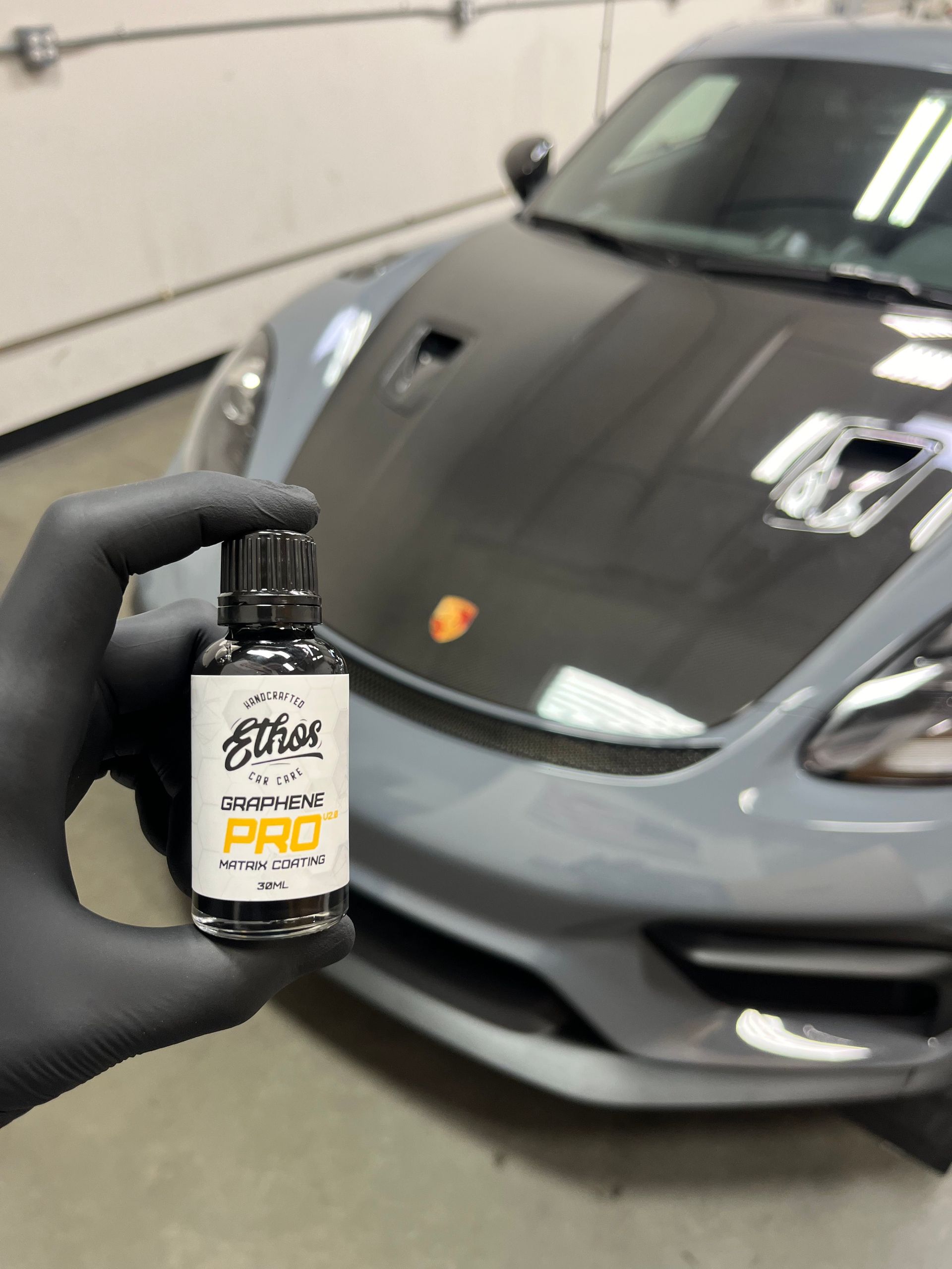 Gloved hand holding a bottle of car detailing product in front of a gray Porsche.