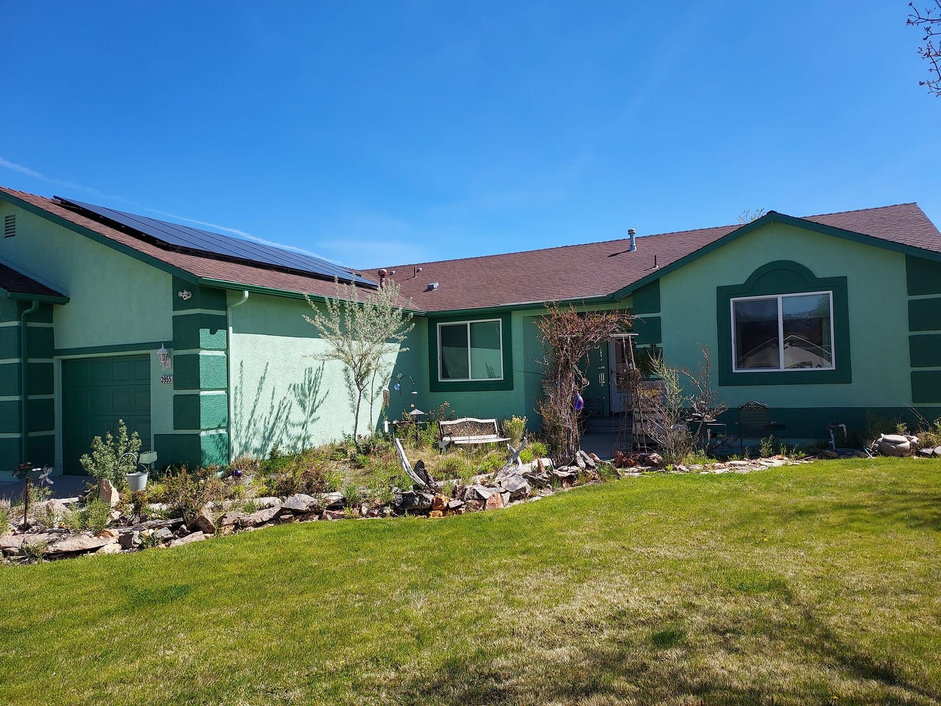 A one-story light green house with dark green trim, solar panels on the roof, and a front yard with rocks and landscaping.