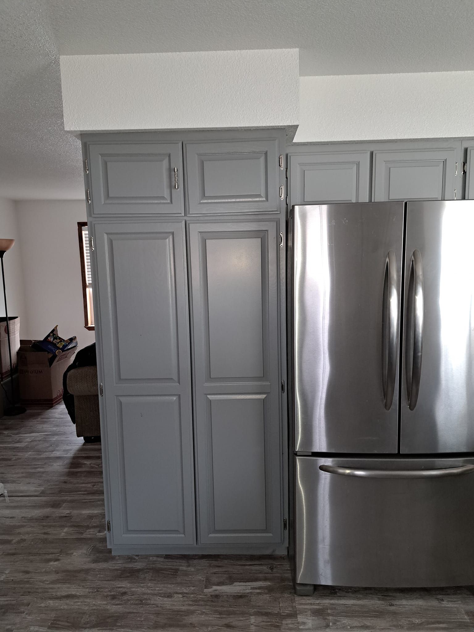 A tall gray cabinet stands next to a stainless steel refrigerator beneath a white textured bulkhead in a kitchen.