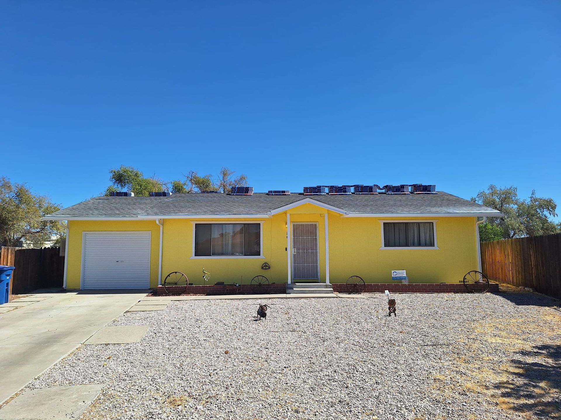 A single-story, bright yellow suburban house with a white garage door, dark roof, and a gravel front yard under a blue sky.