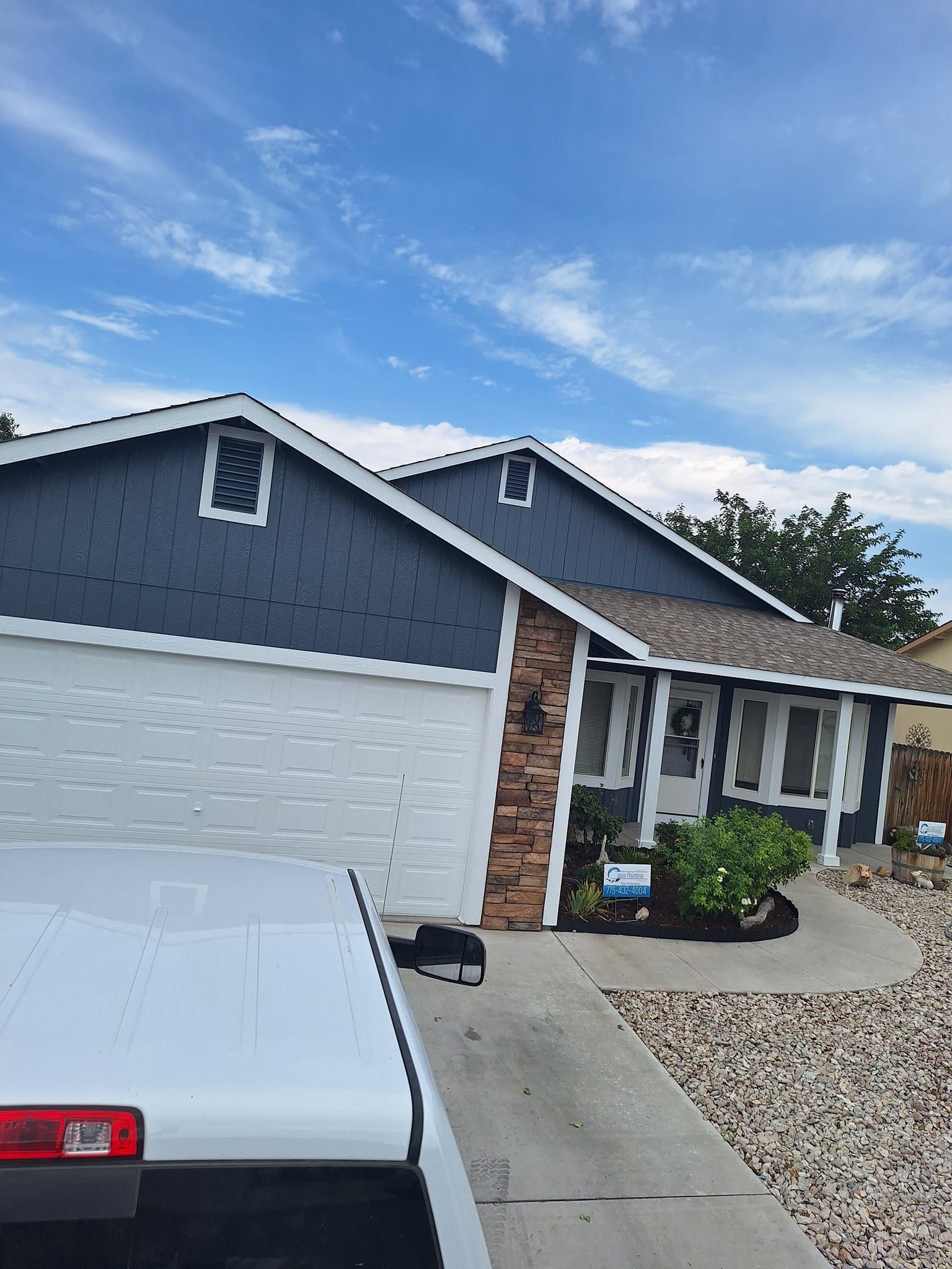 A single-story blue house with a white garage door, stone pillar accent, and a light-colored rock driveway.