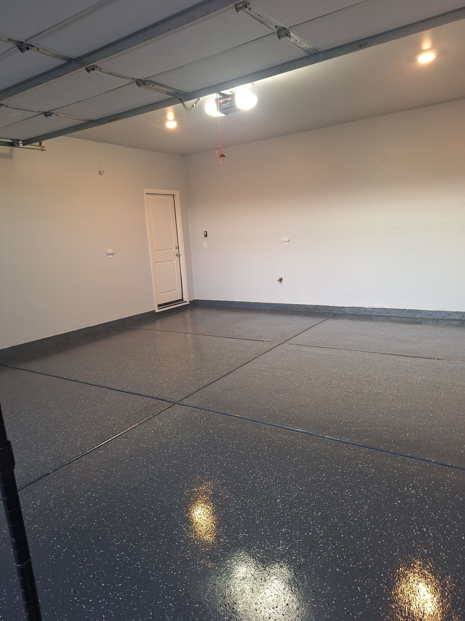 A garage interior with a grey, speckled epoxy floor and plain white walls, featuring an interior door and ceiling lights.