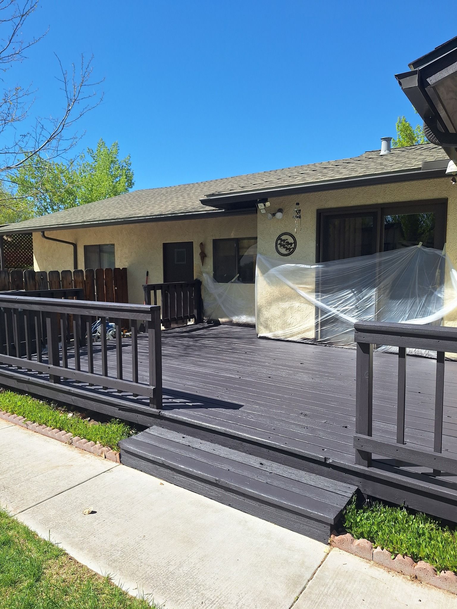 A wide, dark gray wooden deck with railings and steps extends from the back of a light-colored home under a clear blue sky.