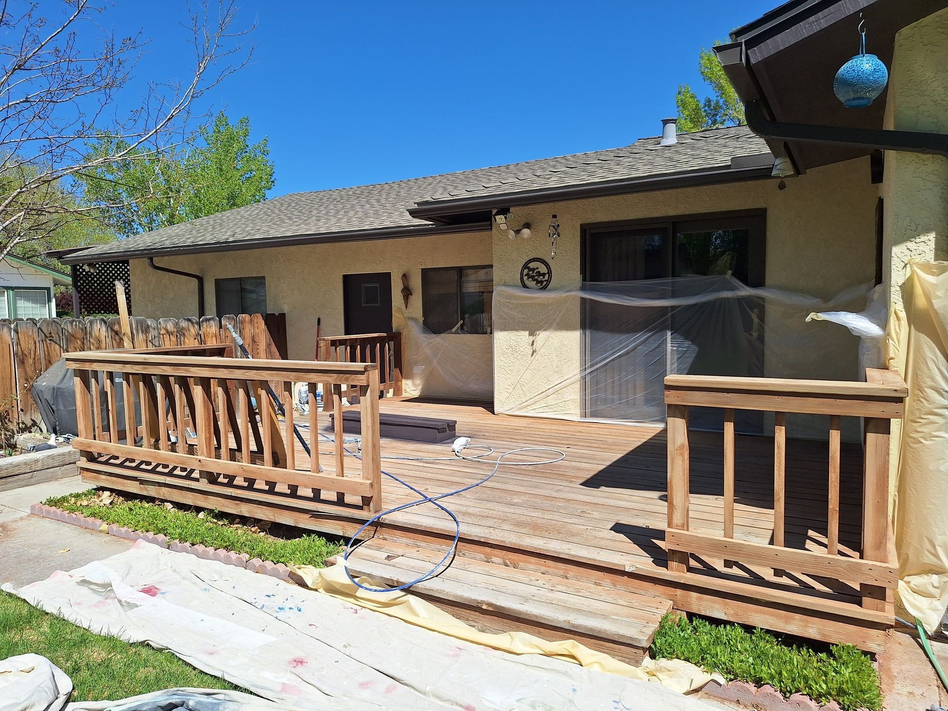 A wooden deck with railings attaches to a cream-colored house under a clear blue sky, with drop cloths on the ground.