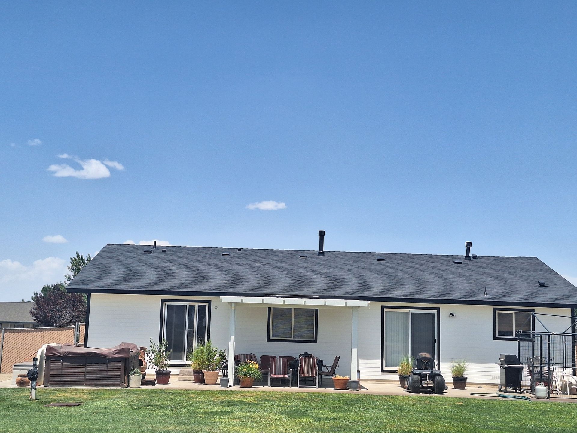 The rear exterior of a single-story white house with a dark shingled roof, a patio with furniture, and a grassy backyard.