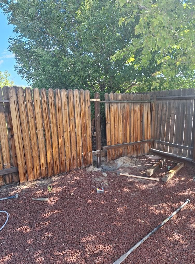 A wooden fence with a missing section between two posts, set against a tree in a gravel yard.