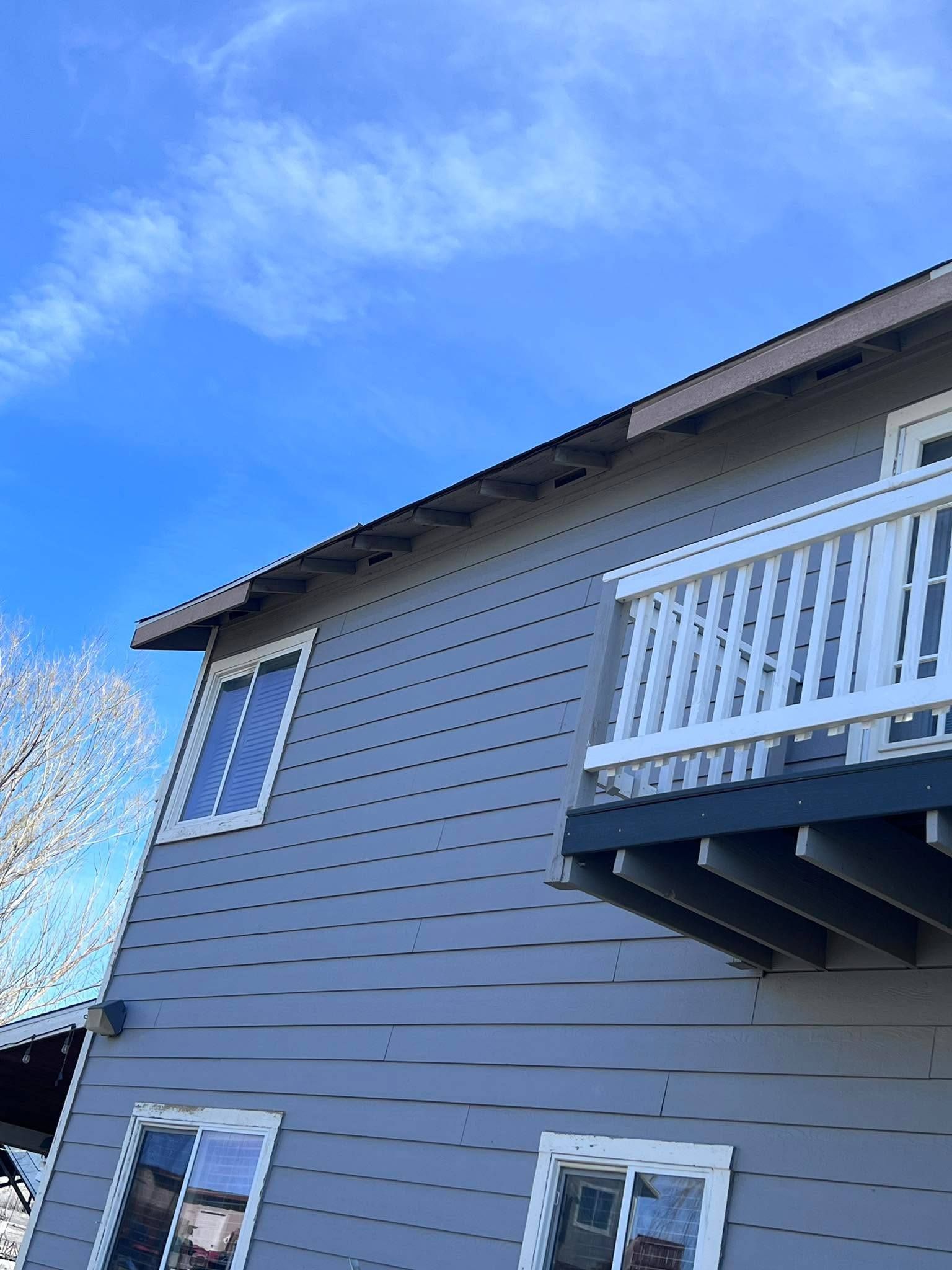 A side view of a gray, multi-story house with horizontal siding, white-framed windows, and an upper-level balcony.