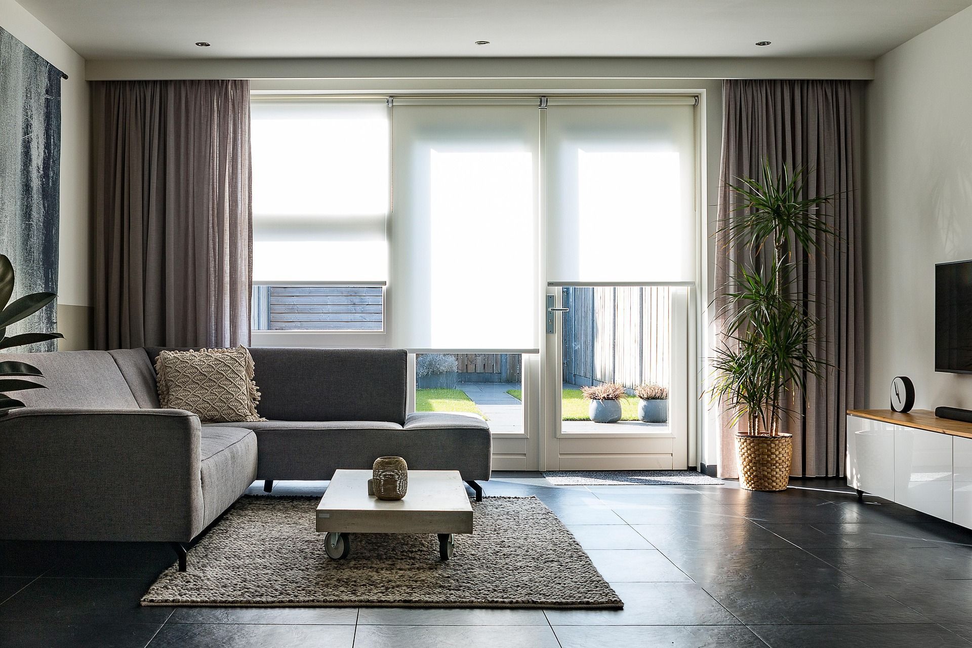 A modern living room with a grey sectional sofa, coffee table, rug, and large windows with curtains and roller shades.