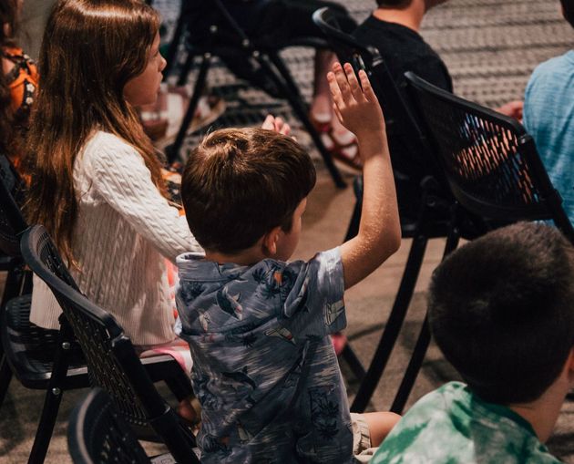 A group of children are sitting in chairs with their hands up.