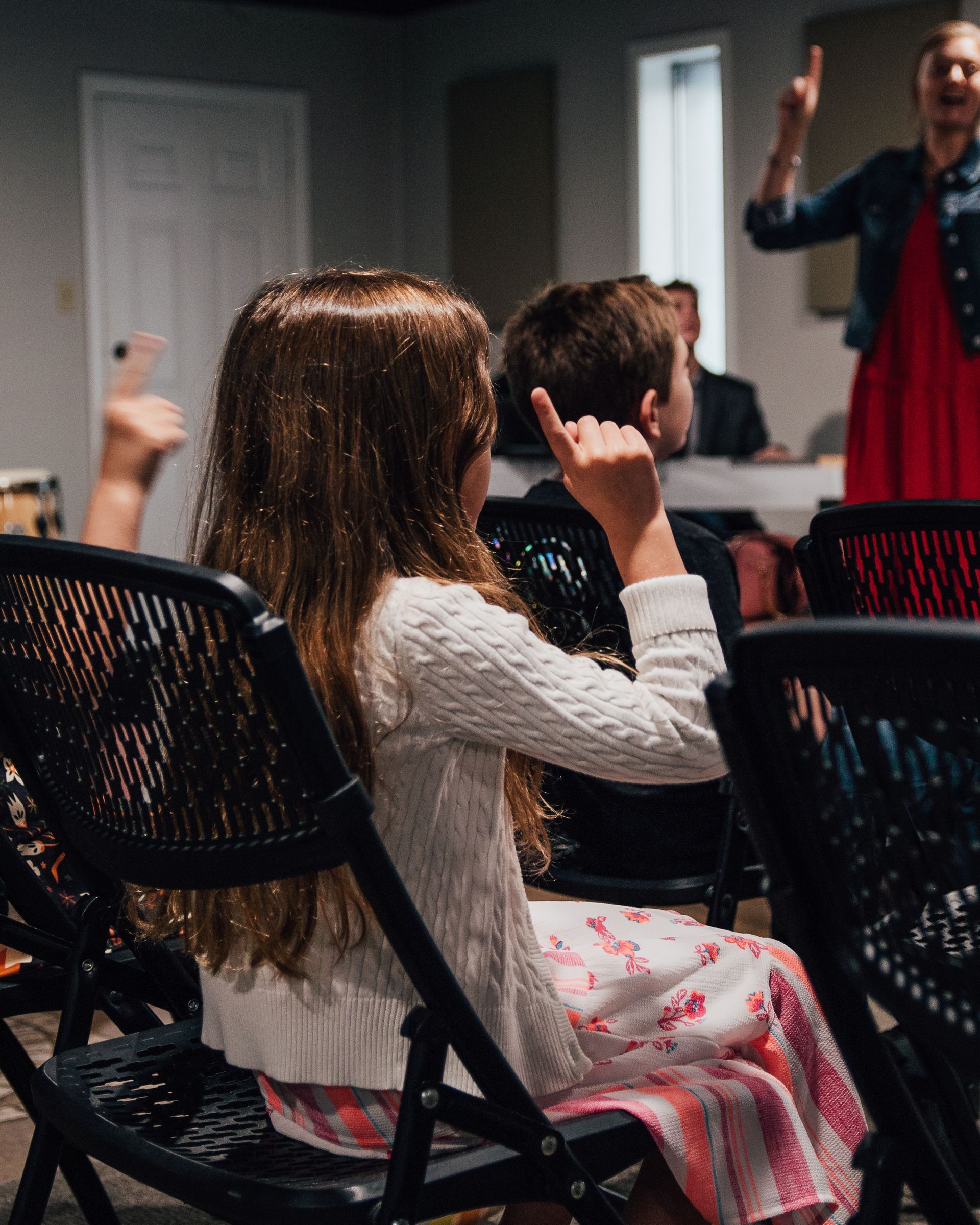 A group of children are sitting in chairs while a woman stands in the background.