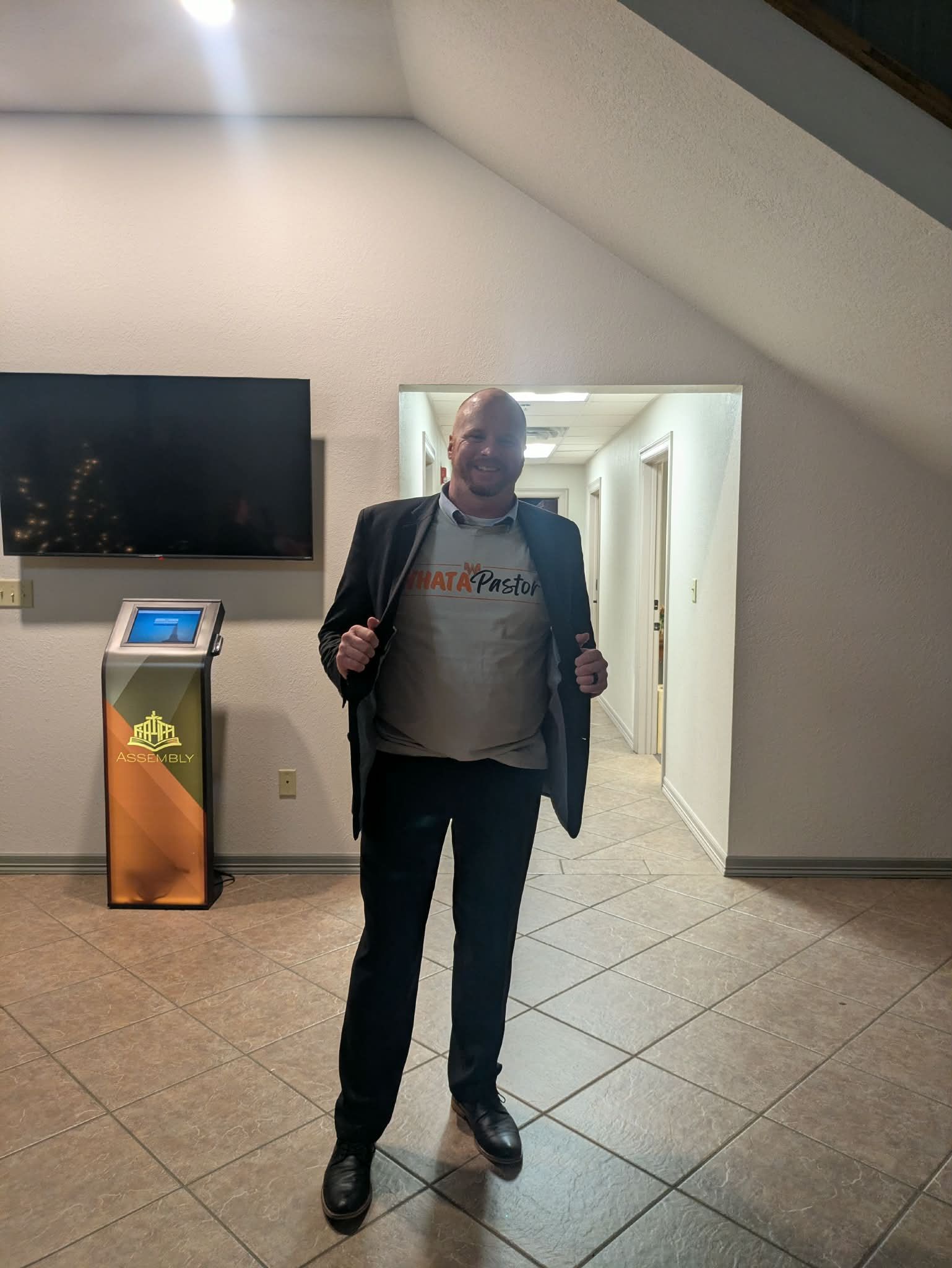 Man in blazer and t-shirt stands indoors with arms open, near a hallway.
