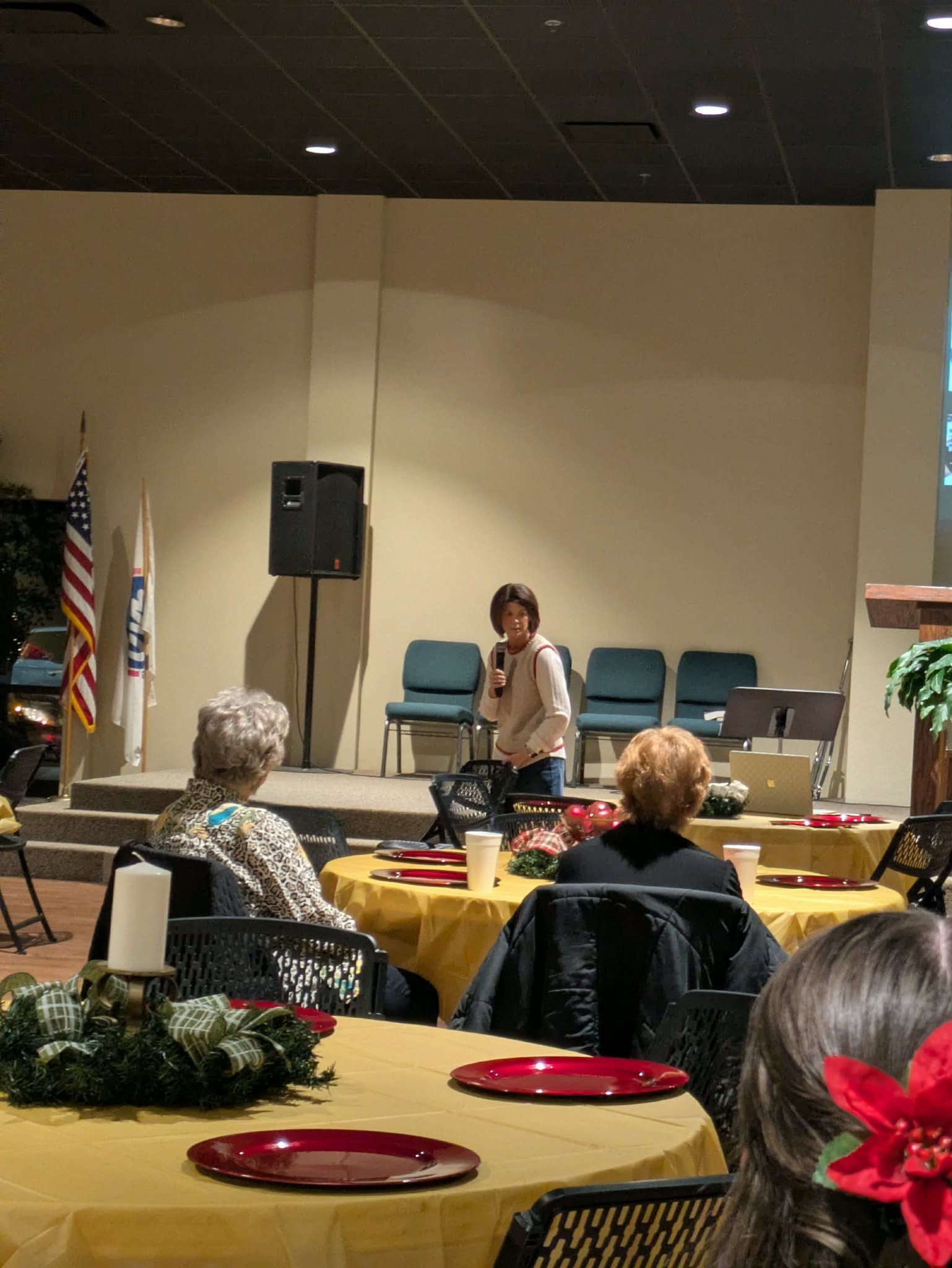 A woman speaking at a small gathering, with attendees seated at round tables.
