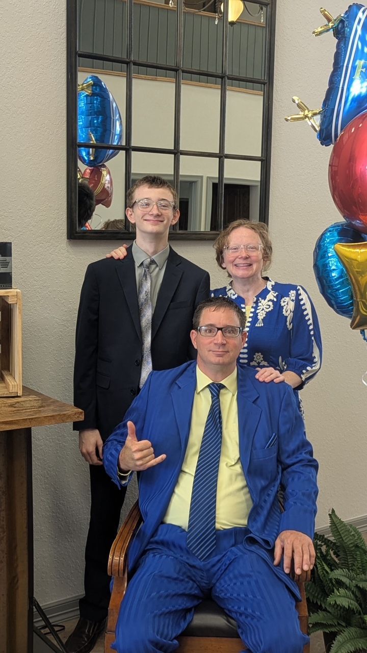 2 people  standing behind a sitting man with colorful balloons beside