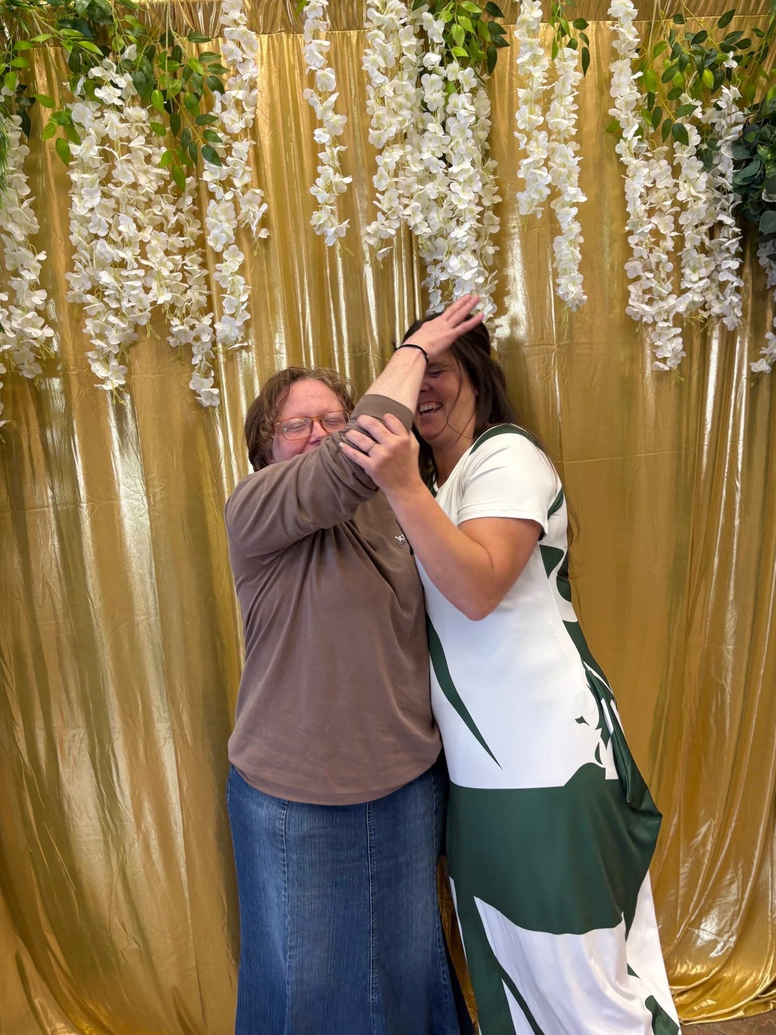Two people posing playfully, one raising the other's arm against a gold fabric backdrop with hanging white florals.
