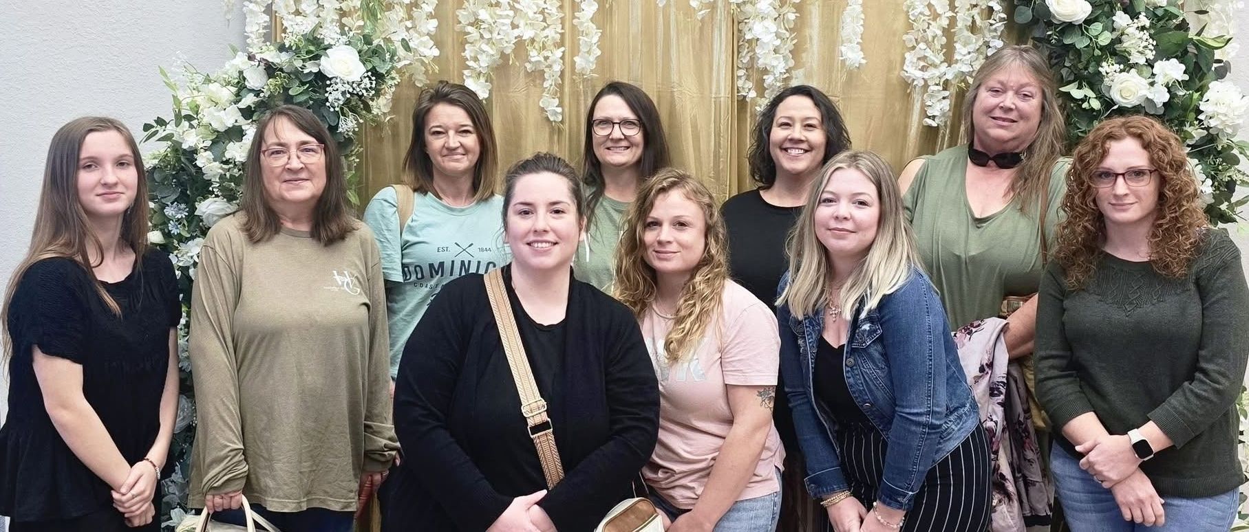 A group of ten people stand smiling in front of a floral backdrop and gold curtain at an indoor event.