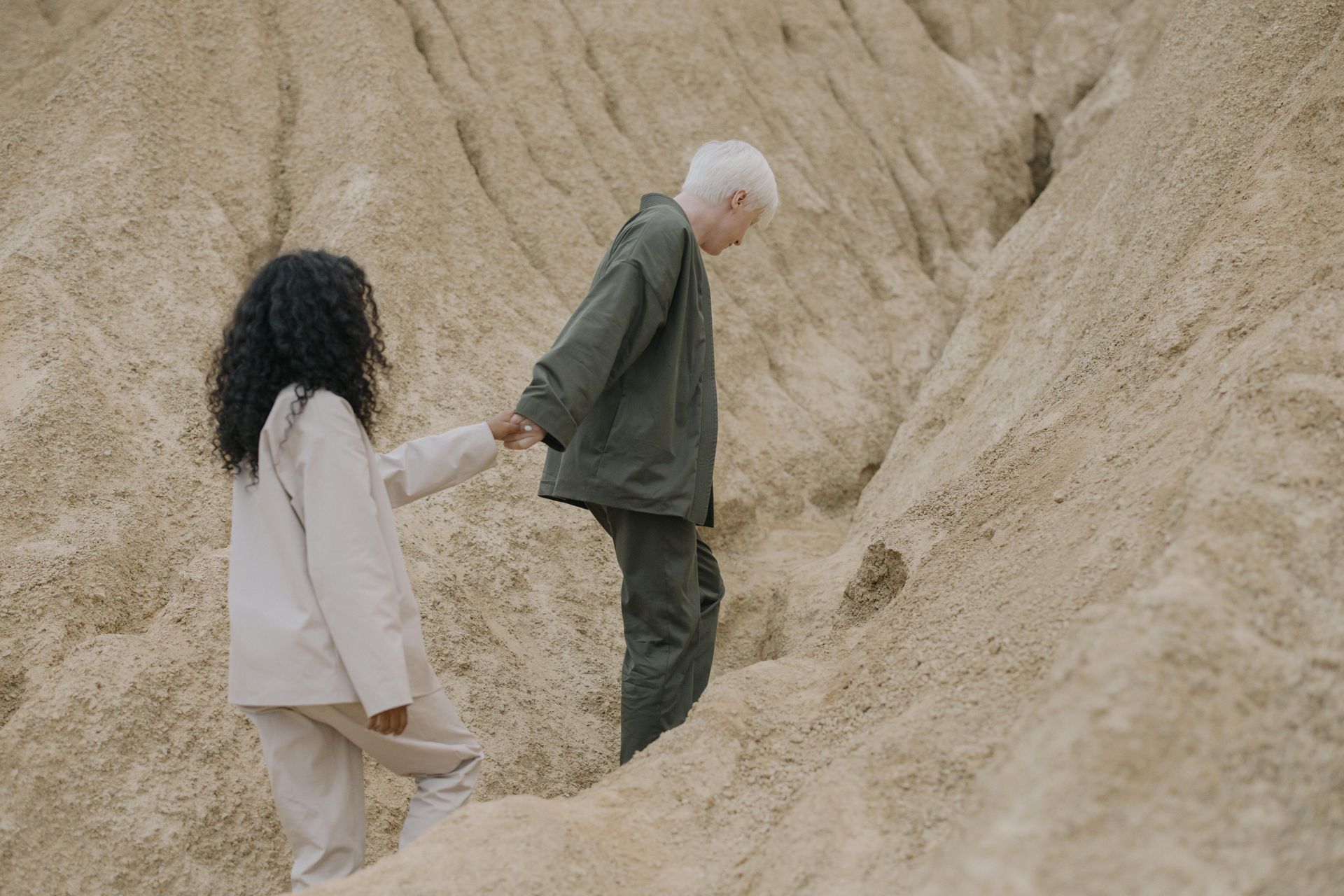Woman helping person climb a sandy hill, holding hands. Both wearing light-colored clothing.