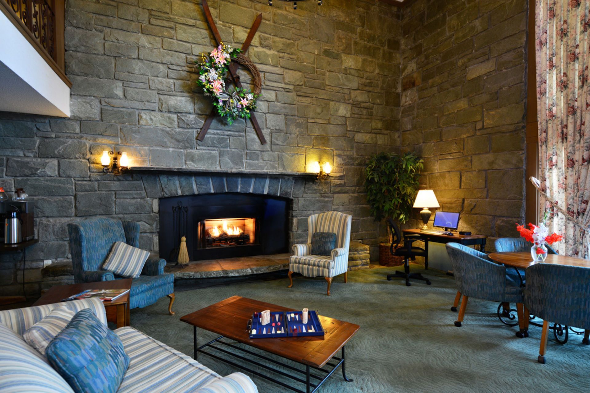 Cozy lodge living room with a stone fireplace, chairs, and a coffee table. A wreath hangs above the fire.