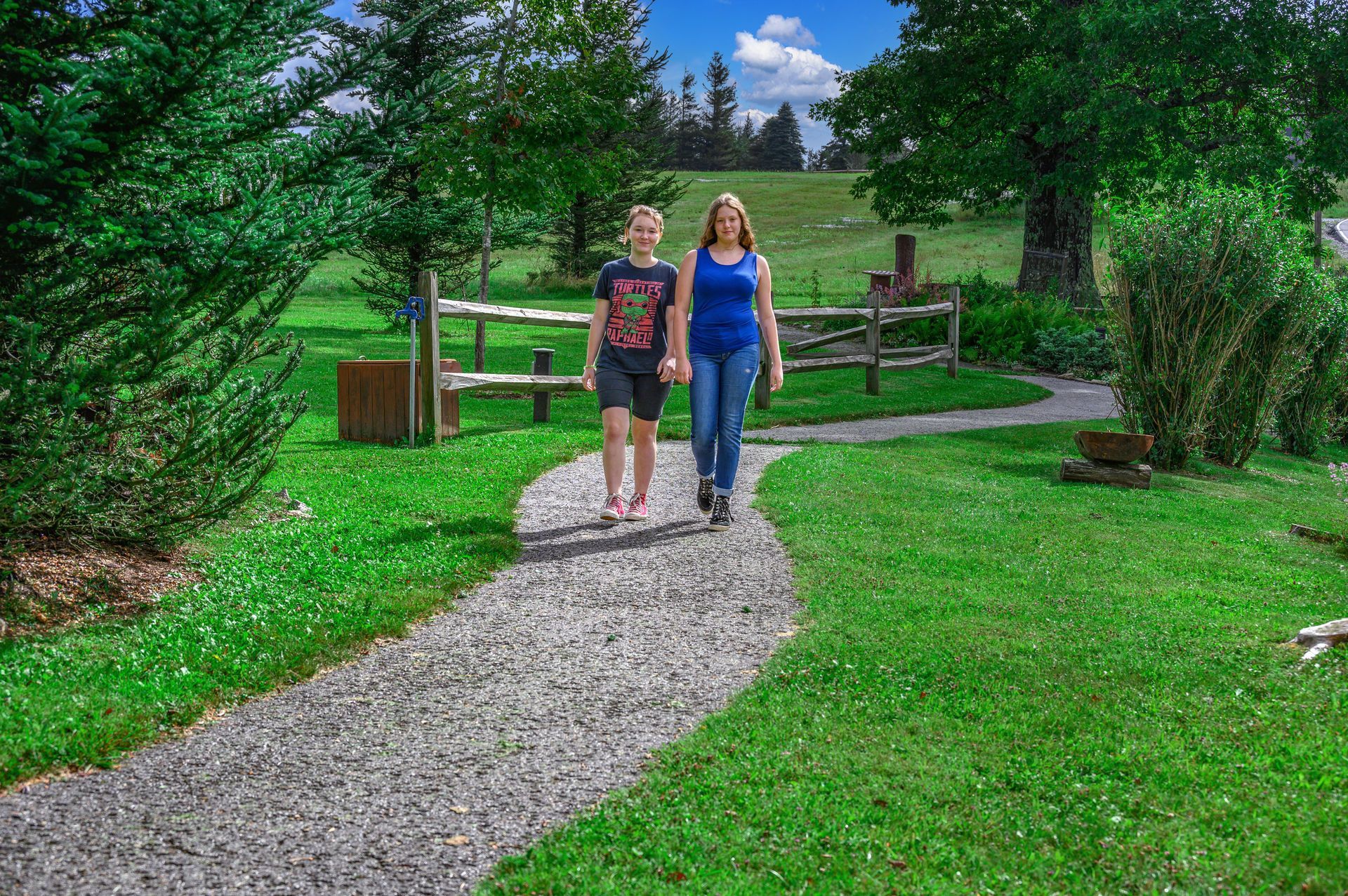 Two people walking along a gravel path in a park. Green grass and trees surround them.