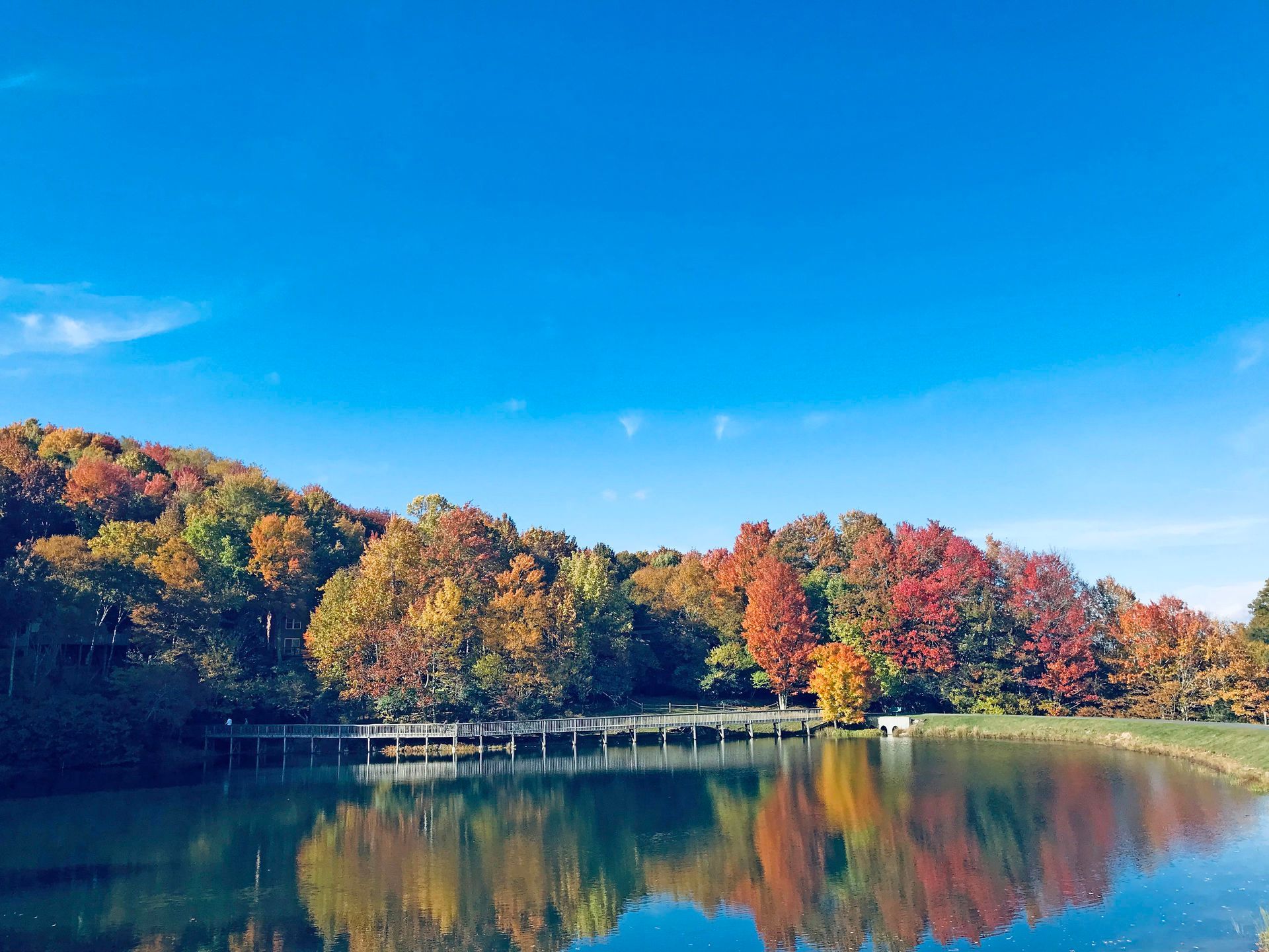 Bright blue sky over a lake reflecting vibrant fall foliage; a wooden bridge extends over the water.