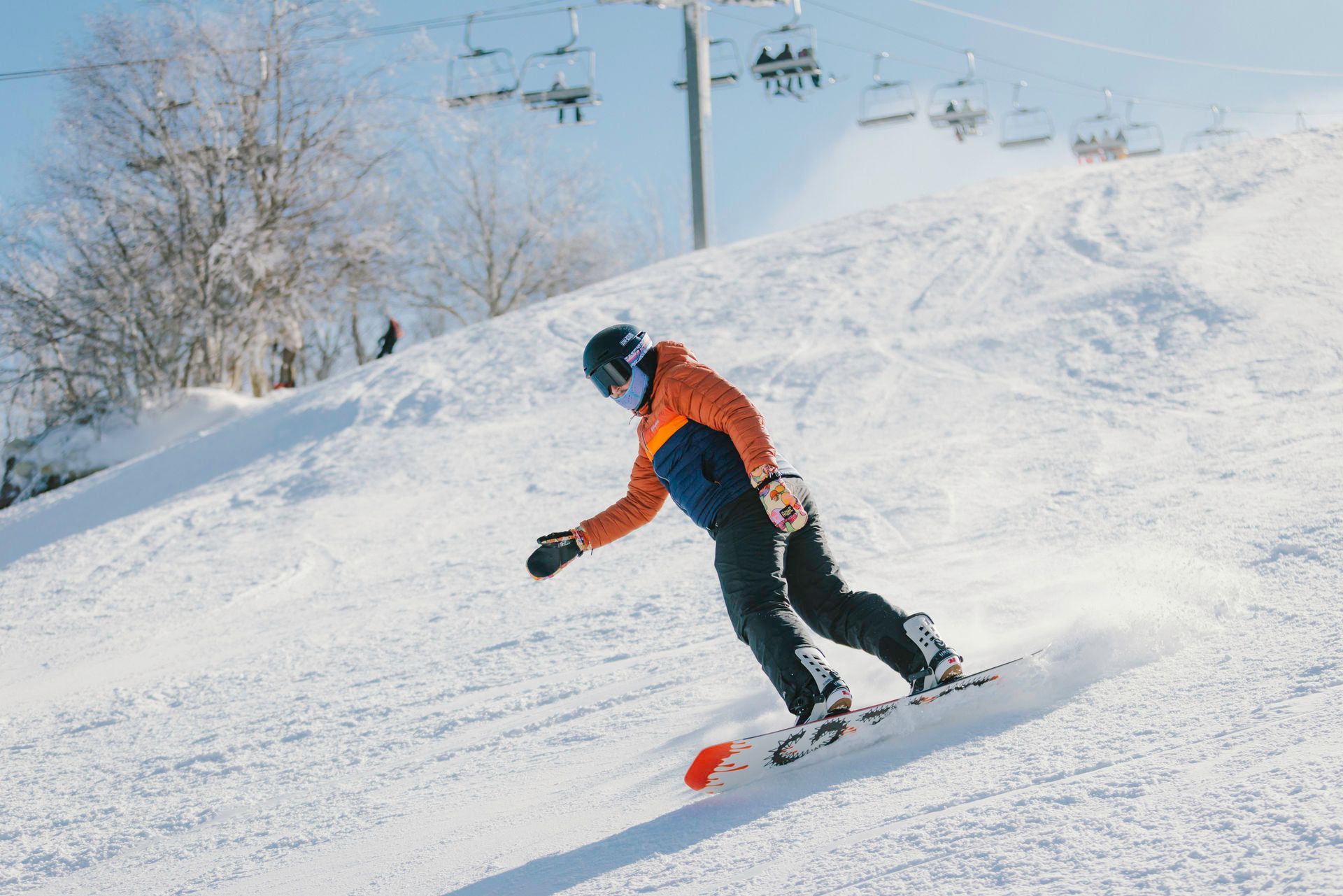 Snowboarder in orange and blue jacket carves down snowy slope under a bright sky.