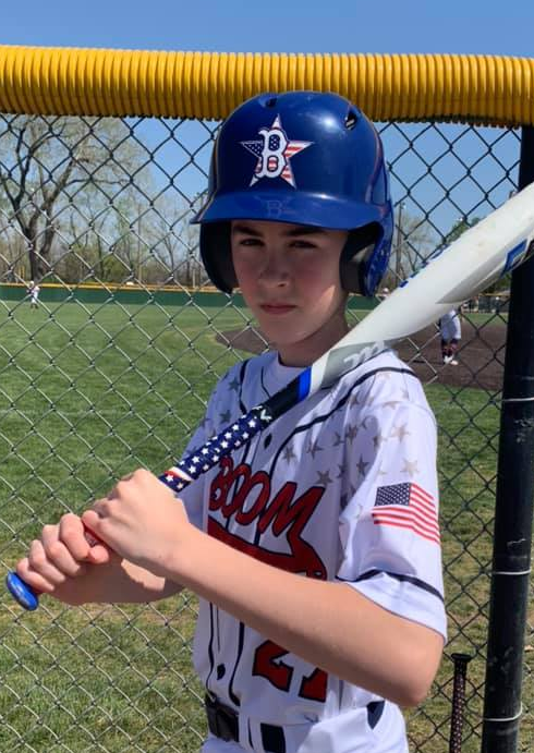 A boy in a baseball uniform, holding a bat, with stickers on his batting helmet