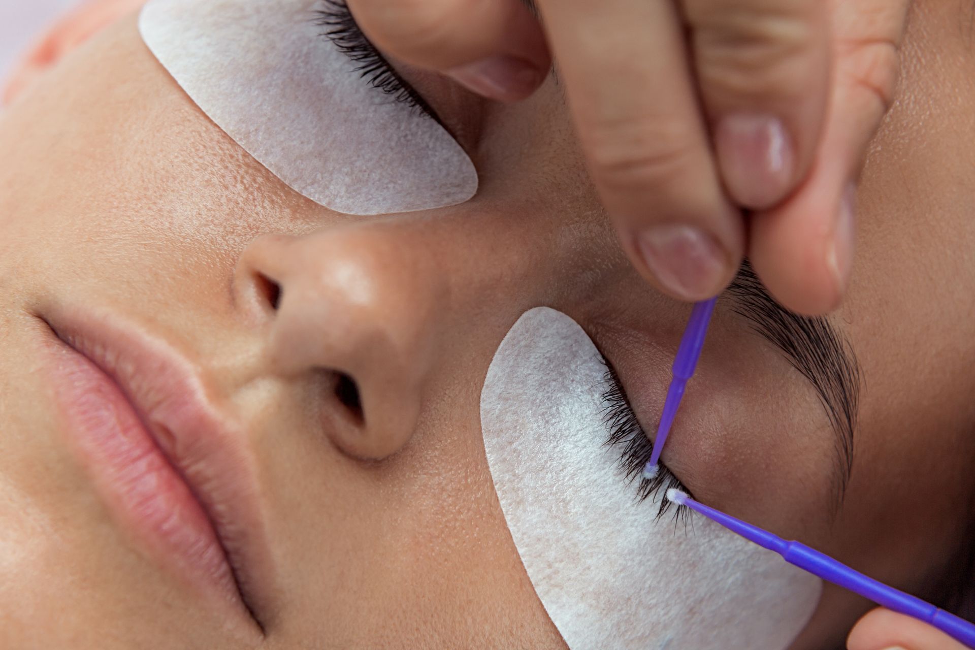 Close-up of a woman receiving eyelash extensions. A technician uses a tool to apply lashes, with eye pads protecting the lower lashes.