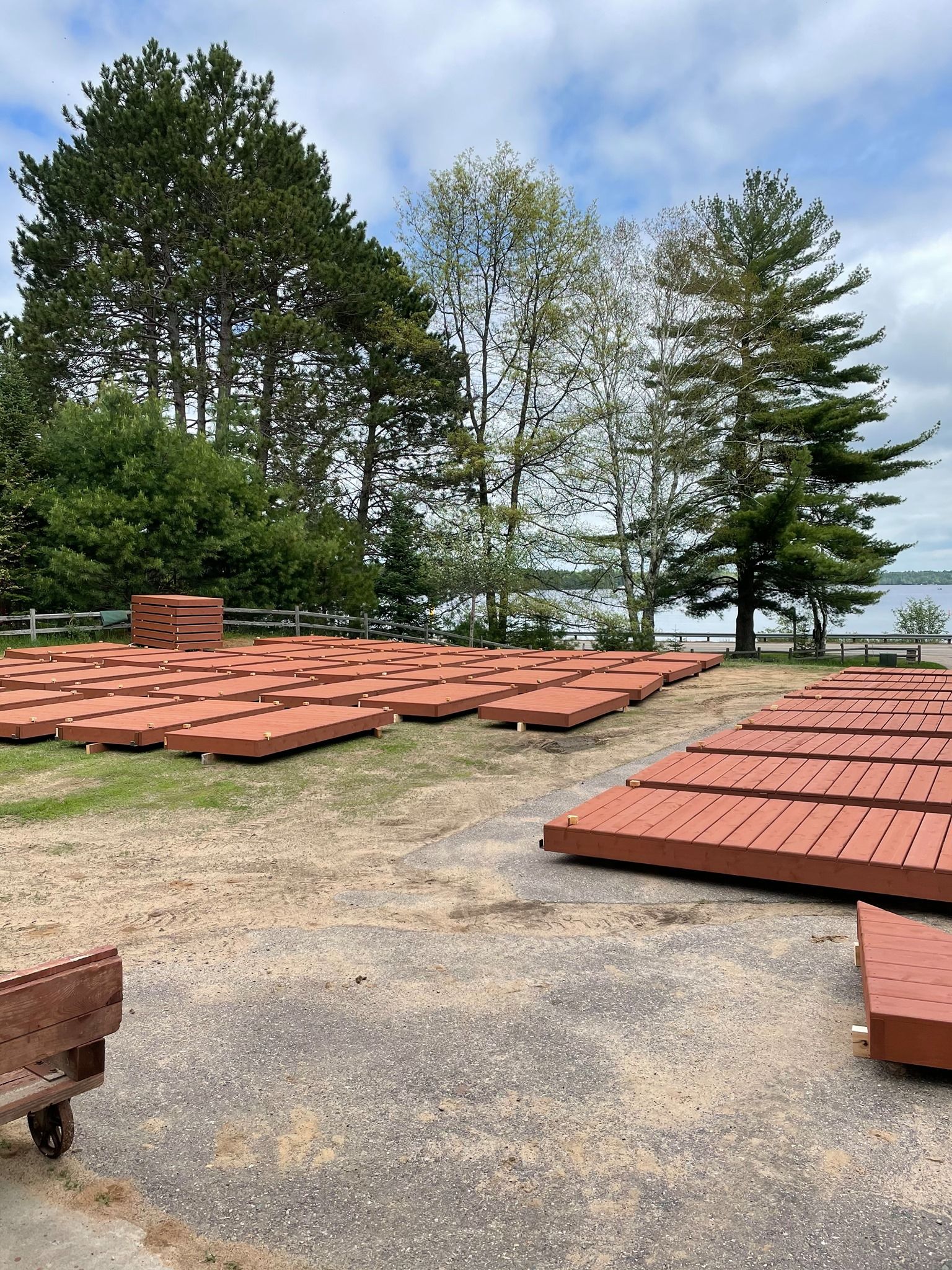 Numerous reddish-brown, rectangular dock sections are laid out on a gravel lot near a body of water and trees.
