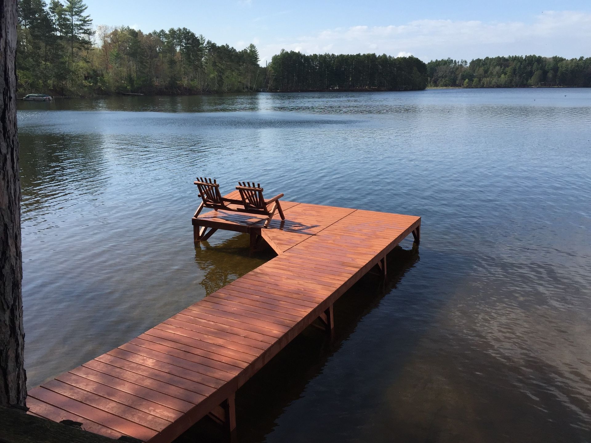 Two brown Adirondack chairs sit on a reddish wooden L-shaped dock extending into a calm lake surrounded by trees.