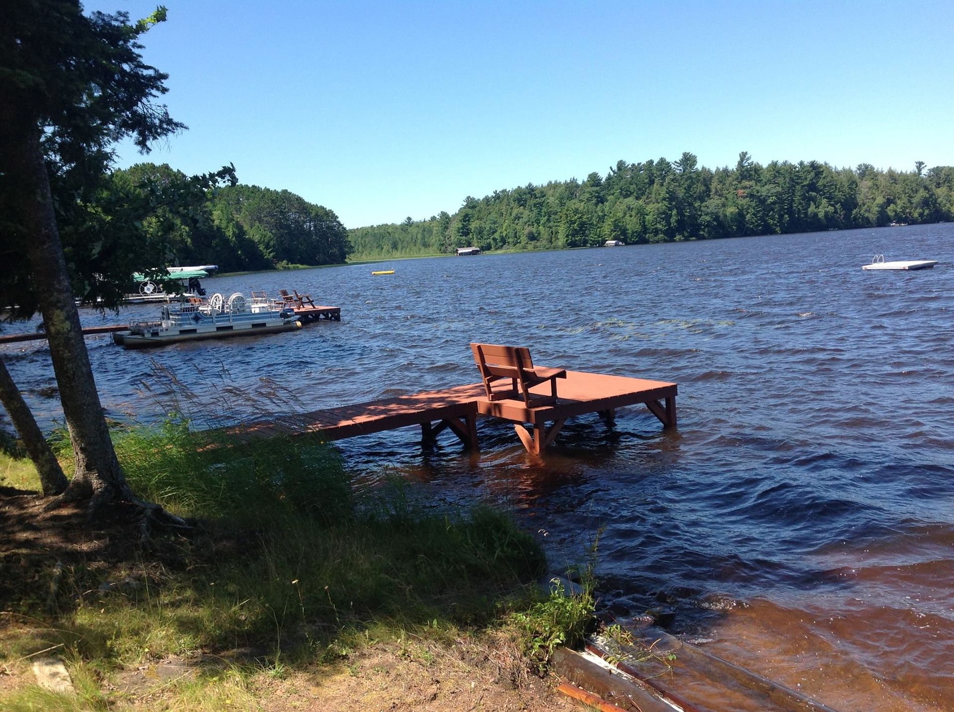 A wooden dock with a bench sits in a sunlit lake, with a boat moored nearby and trees lining the distant shore.