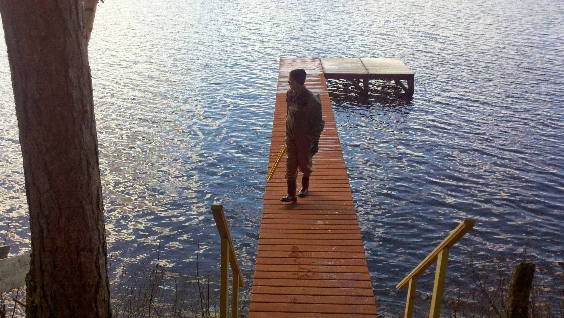 A person stands on a long, orange-painted wooden dock extending into a sparkling blue lake.