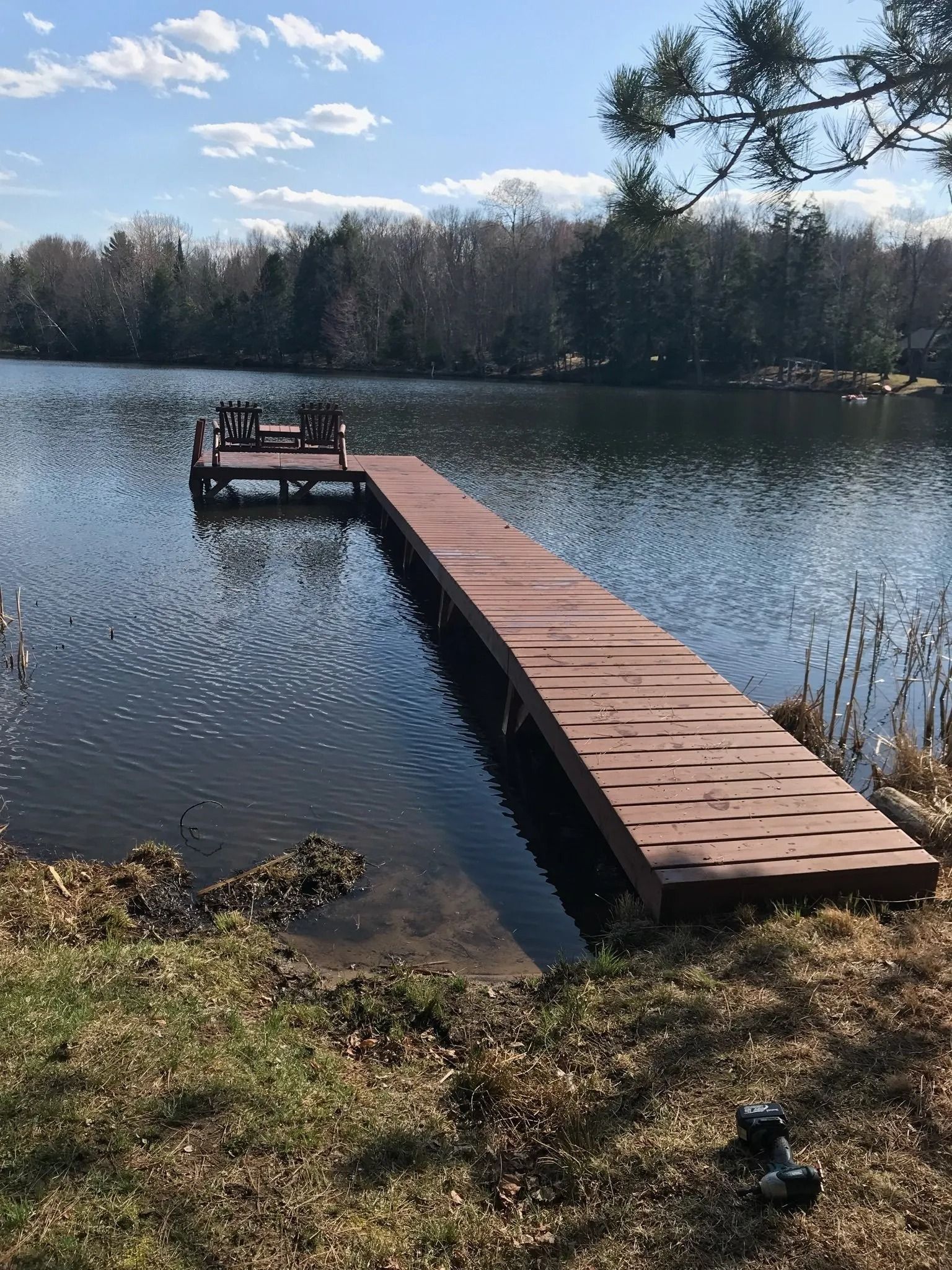 A long, brown wooden dock extends into a calm, blue lake surrounded by trees under a sunny sky.