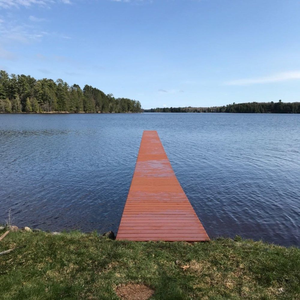 A wooden pier extends into a calm, blue lake, flanked by a treelined shore under a clear, sunny sky.