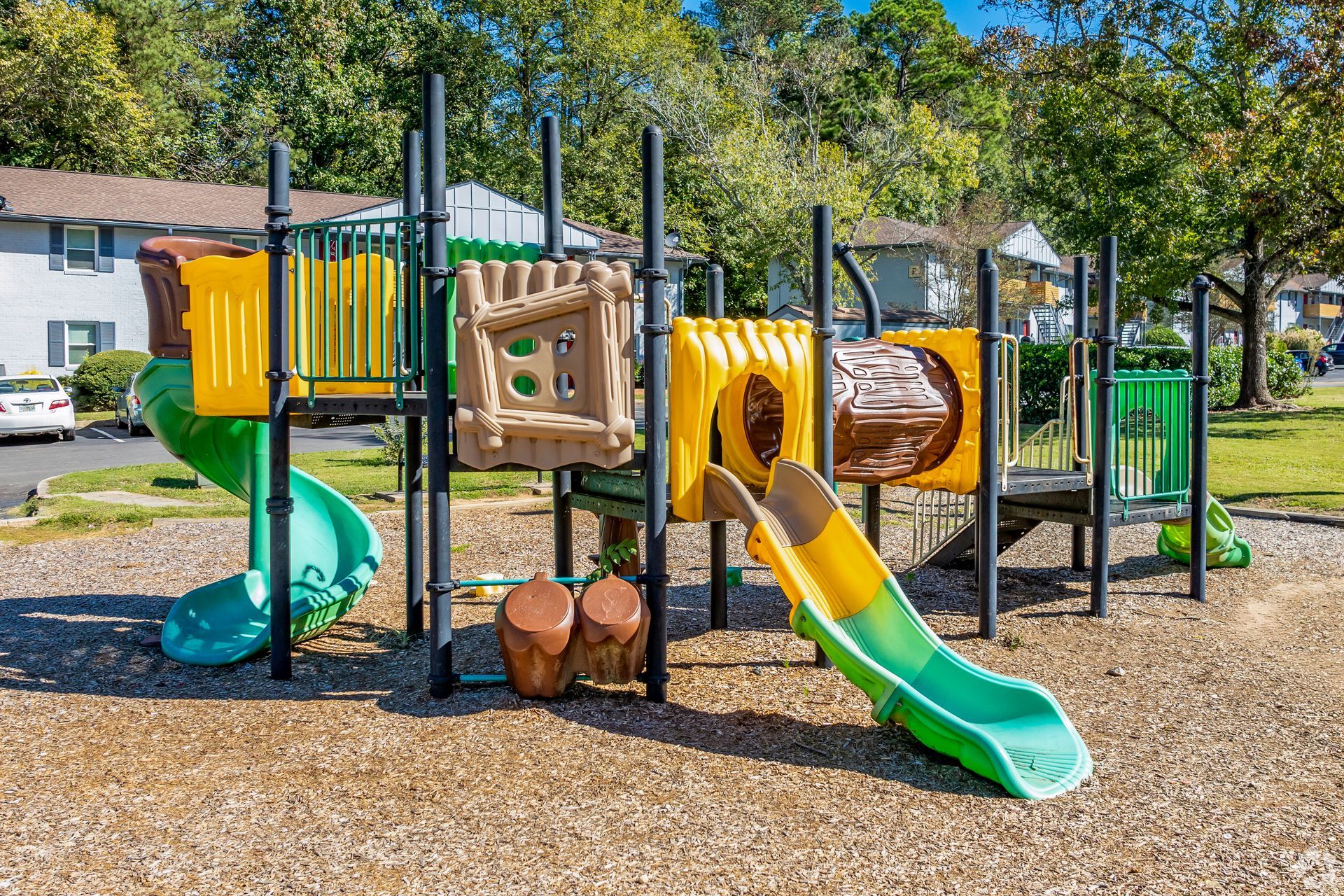 Playground with slides, climbing structures, and mulch ground cover in a park setting.