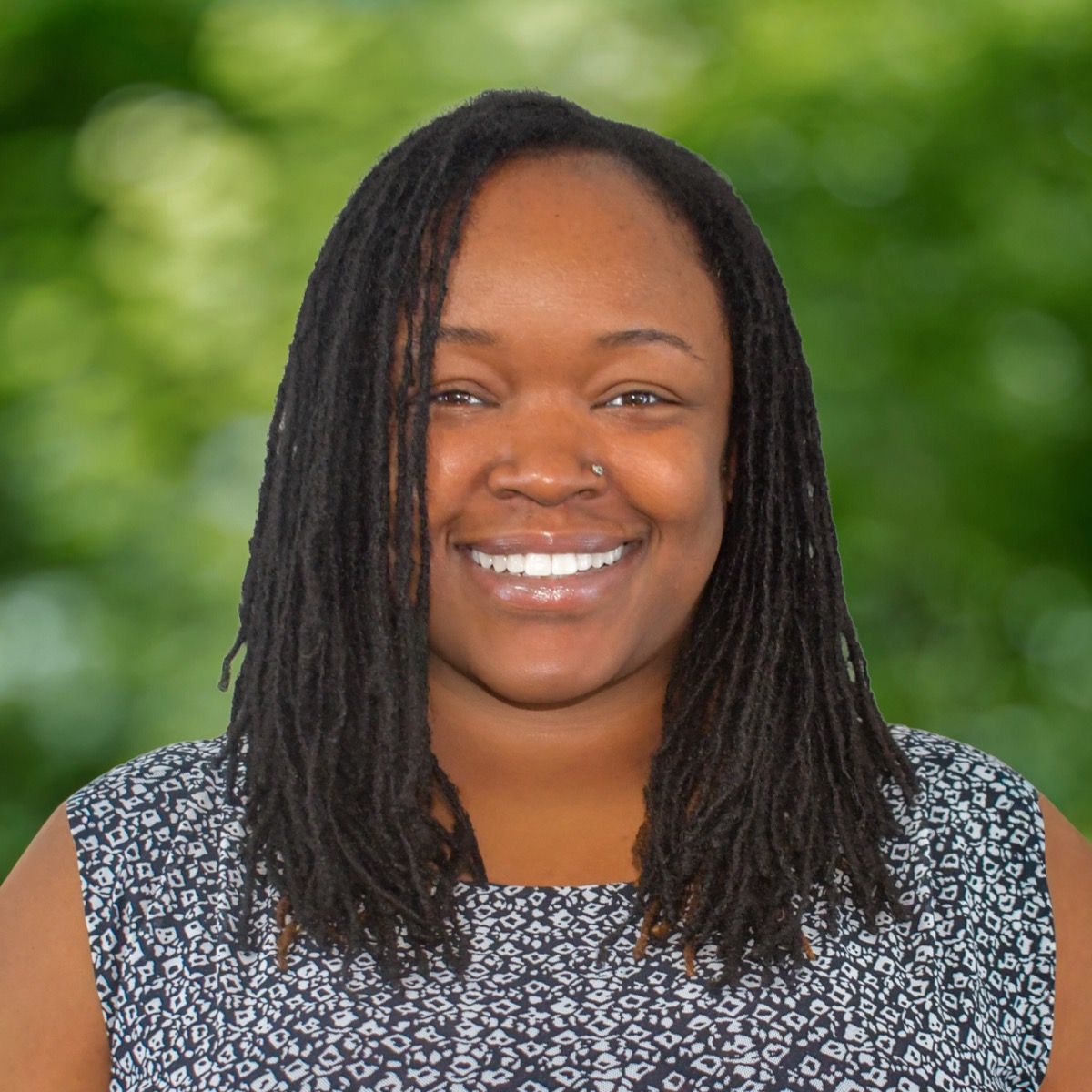 Smiling man with fair skin and a beard in a blue polo shirt, outdoors with green foliage.