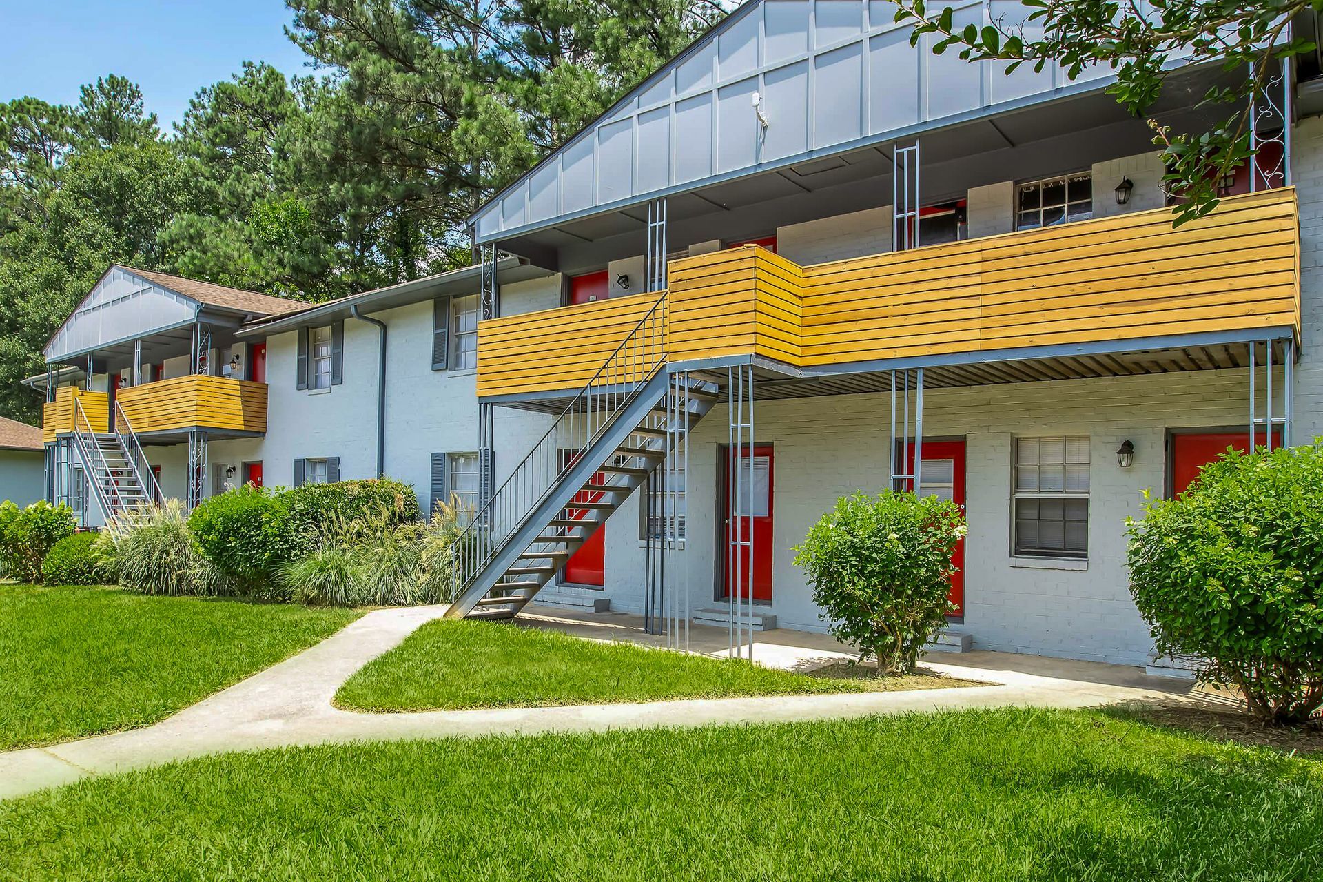Apartment building exterior, white with red doors, yellow roof, metal stairs, green lawn and bushes.